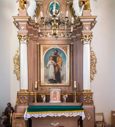 Zdjęcie nr 2: Obraz w kształcie stojącego prostokąta zamkniętego łukiem półkolistym w srebrnej, dekorowanej ornamentami ramie. W centrum kompozycji znajduje się całopostaciowe przedstawienie św. Józefa spoglądającego na stojącego przed nim młodego Jezusa. Jezus jest ustawiony frontalnie, w kontrapoście, patrząc na widza, prawą rękę wyciąga przed siebie, lewą wskazuje znajdujące się na jego piersi gorejące serce otoczone koroną cierniową i zwieńczone krzyżem. Jezus ma owalną twarz, szeroko otwarte, ciemne oczy, wąski nos  i małe usta. Jego blond, falowane, krótkie włosy są rozdzielone przedziałkiem. Jest ubrany w długą, białą tunikę przewiązaną w pasie niebieskim paskiem. Jezus stoi na globie ziemskim oplecionym wicią z białymi kwiatami. Ustawiony za nim Józef w lewej ręce trzyma kwiat lilii, ma drobną twarz okoloną ciemnymi włosami i sięgającą dekoltu brodę. Jest ubrany w ciemnobrązową tunikę wykończoną pod szyją złotą  lamówką z kamieniami szlachetnymi. Na to ma narzucony żółty płaszcz z niebieskim podbiciem, który zakrywa ręce, łamiąc się grubymi fałdami i przechodzi z przodu poniżej pasa. Obie postacie mają nad głowami wąskie, okrągłe nimby; nimb Jezusa jest wypełniony krzyżem. Tło przedstawienia stanowi niebieskie niebo przesłonięte w dolnej części białymi obłokami.  W prawym dolnym rogu sygnatura „Malował Władysław Rossowski”, poniżej umieszczona jest częściowo przesłonięta data. 