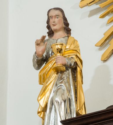 Zdjęcie nr 1: Figura pełna na cokole. Święty Jan ukazany w całej postaci, stojący frontalnie, w kontrapoście, z lewą nogą wysuniętą do przodu. W lewej ręce trzyma kielich, a prawą czyni nad nim gest błogosławieństwa; patrzy przed siebie. Święty ma młodzieńczą, podłużną twarz z asymetrycznie ustawionymi oczami, prostym nosem i wąskimi, silnie zarysowanymi ustami. Długie, ciemnobrązowe włosy zwinięte w pukle opadają mu na plecy. Ubrany jest w srebrną suknię, która fałduje się nad zgiętym kolanem. Na plecy i lewe ramię ma zarzucony złoty płaszcz. Polichromia w odsłoniętych partiach ciała traktowana naturalistycznie.