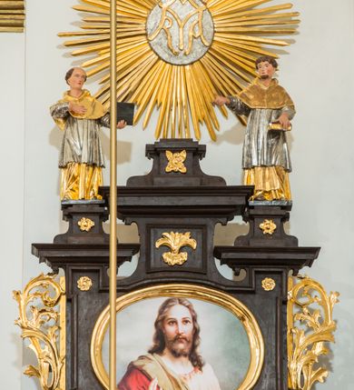 Zdjęcie nr 2: Ołtarz architektoniczny, przyścienny, jednoosiowy, dwukondygnacyjny ze zwieńczeniem. Nastawa umieszczona jest na skrzyniowej mensie, której antepedium dekoruje ciemnoróżowa tkanina zdobiona wzorem zwiniętej wici roślinnej. Cokół jest wypełniony w centrum prostokątną płyciną, a na osiach zewnętrznych plakietami ornamentalnymi utworzonymi z motywów roślinnych z dziecięcymi główkami. Nastawa flankowana ustawionymi na wysuniętych partiach cokołu kolumnami dźwigającymi fragmenty pełnego belkowania, które podtrzymują partie urwanego przyczółka. W polu centralnym nisza w kształcie stojącego prostokąta z uskokiem, ponad którym gzyms i łuk półokrągły. W niszy umieszczono ujęty złoconą, ażurową ramą obraz z wizerunkiem Matki Boskiej Częstochowskiej. Powyżej obrazu, w łuku, plakieta dekoracyjna. Druga kondygnacja w kształcie leżącego prostokąta z uszakami jest zwieńczona trzema postumentami, z których wszystkie dźwigają fragmenty gzymsu podtrzymującego cokoły. Na zewnętrznych ustawiono postacie niezidentyfikowanych świętych. Kondygnację ujmują uszaki utworzone z suchej wici akantowej, pole wypełnia owalny obraz z popiersiem błogosławiącego Jezusa. Nastawę wieńczy umieszczony na ścianie monogram maryjny ujęty w kształt kropi i umieszczony na tle glorii promienistej. 
Struktura polichromowana na kolor ciemnobrązowy ze złoconymi kolumnami oraz złoconą i srebrzoną ornamentyką oraz detalami architektonicznymi.
