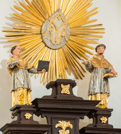 Zdjęcie nr 5: Ołtarz architektoniczny, przyścienny, jednoosiowy, dwukondygnacyjny ze zwieńczeniem. Nastawa umieszczona jest na skrzyniowej mensie, której antepedium dekoruje ciemnoróżowa tkanina zdobiona wzorem zwiniętej wici roślinnej. Cokół jest wypełniony w centrum prostokątną płyciną, a na osiach zewnętrznych plakietami ornamentalnymi utworzonymi z motywów roślinnych z dziecięcymi główkami. Nastawa flankowana ustawionymi na wysuniętych partiach cokołu kolumnami dźwigającymi fragmenty pełnego belkowania, które podtrzymują partie urwanego przyczółka. W polu centralnym nisza w kształcie stojącego prostokąta z uskokiem, ponad którym gzyms i łuk półokrągły. W niszy umieszczono ujęty złoconą, ażurową ramą obraz z wizerunkiem Matki Boskiej Częstochowskiej. Powyżej obrazu, w łuku, plakieta dekoracyjna. Druga kondygnacja w kształcie leżącego prostokąta z uszakami jest zwieńczona trzema postumentami, z których wszystkie dźwigają fragmenty gzymsu podtrzymującego cokoły. Na zewnętrznych ustawiono postacie niezidentyfikowanych świętych. Kondygnację ujmują uszaki utworzone z suchej wici akantowej, pole wypełnia owalny obraz z popiersiem błogosławiącego Jezusa. Nastawę wieńczy umieszczony na ścianie monogram maryjny ujęty w kształt kropi i umieszczony na tle glorii promienistej. 
Struktura polichromowana na kolor ciemnobrązowy ze złoconymi kolumnami oraz złoconą i srebrzoną ornamentyką oraz detalami architektonicznymi.
