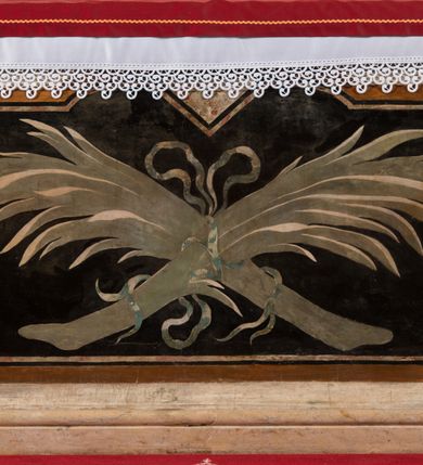 Zdjęcie nr 25: Na niskim postumencie o krawędzi przedniej wyciętej linią falistą osadzony ołtarz w kształcie wybrzuszonej górą tumby. Jego antepedium wypełnione marmoryzacją (w technice skaliola) z krzyżującymi się palmami męczeństwa. Po bokach ołtarza dwa ukośnie dostawione postumenty, na których dwie figury aniołów flankujących owalny obraz w złoconej ramie z przedstawieniem męczeństwa św. Sebastiana. Malowidło jest obwiedzione dekoracją sztukatorską w formie obłoków z główkami lub postaciami aniołów, zwieńczone krzyżem i glorią promienistą. Anioły klęczące na postumentach ukazane są w poruszonych pozach, w obficie drapowanych szatach, tworzących grube, mięsiste poły. Anioł po lewej stronie ma głowę zwróconą w kierunku obrazu, a ręce rozkłada na boki, anioł po przeciwnej stronie głowę odwraca w odwrotnym niż obraz kierunku, a jego ręce są uniesione na wysokość klatki piersiowej i skierowane do środka. Nad obrazem anioł po lewej stronie niemalże leży na ramie obrazu, w lewej ręce trzyma krzyż, prawą lekko unosi. Ubrany jest w miękko drapowane szaty, odsłaniające dolną część jego prawej nogi. Duże skrzydła o gęstych piórach rozłożone są niesymetrycznie. Po przeciwnej stronie ukazane putta i główki modelowanych sumarycznie, pulchnych aniołków. Dolne partie wykonane z kamienia, marmoryzowane w jasnych kolorach z czarnym polem antepedium wypełnionym jasnozielonymi palmami męczeństwa. Partie rzeźbiarskie białe, rama obrazu, krzyż i gloria złocone. 

