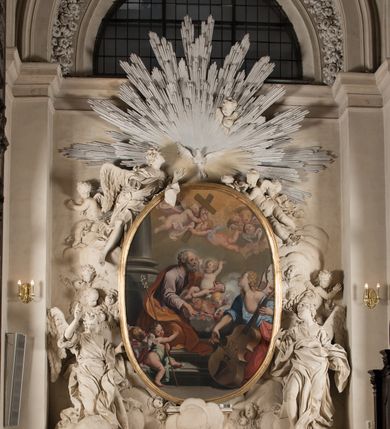 Zdjęcie nr 2: Na niskim postumencie o krawędzi przedniej zarysowanej linią falistą osadzony ołtarz w kształcie tumby, wybrzuszonej w górnej partii. Jego antepedium wypełnione marmoryzacją z owalnym, pustym polem i pękami różnokolorowych kwiatów. Po bokach ołtarza dwa ukośnie dostawione kamienne postumenty, na których dwie figury aniołów flankujących owalny obraz w złoconej ramie z przedstawieniem św. Józefa z Jezusem i grającym na kontrabasie aniołem. Malowidło obwiedzione dekoracją sztukatorską w formie obłoków z główkami lub postaciami aniołów i zwieńczone glorią promienistą z gołębicą Ducha Świętego. Anioły klęczące na postumentach ukazane są w poruszonych pozach, w obficie drapowanych szatach, tworzących grube, mięsiste poły, ze skrzydłami o gęstych piórach. Anioł po lewej stronie jest ukazany frontalnie, klęcząc na chmurze na prawym kolanie, z lewą noga wyprostowaną, z głową w trzech czwartych, zwróconą w swoją prawą stronę. Lewą ręką przytrzymuje ramę obrazu, prawą unosi w geście błogosławieństwa. Anioł po prawej stronie głowę kieruje ku obrazowi, jego ręce są uniesione na wysokości klatki piersiowej, dłoń lewą opuszcza, a prawą unosi. Nad górną częścią obrazu został ukazany anioł w pozycji niemalże leżącej, z rozłożonymi rękami, odziany w bogato drapowane szaty odsłaniające dolną część nogi. Duże skrzydła o gęstych piórach rozłożone są niesymetrycznie. Po przeciwnej stronie leży na chmurach nagie putto wśród modelowanych sumarycznie główek anielskich. Dolne partie wykonane z kamienia inkrustowanego czarnym marmurem i marmoryzacją z różnokolorowymi pękami kwiatów, w jasnych kolorach z czarnym polem antepedium z różnobarwnymi kwiatami, partie rzeźbiarskie białe, rama obrazu złocona, gloria bladoniebieska. 