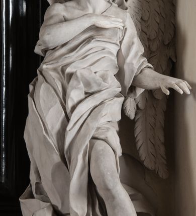 Zdjęcie nr 15: Na niskim postumencie, na rzucie prostokąta z łukiem nadwieszonym od przodu osadzony ołtarz prostopadłościenny. Antepedium marmoryzowane z owalnym polem i rytym przedstawieniem głowy św. Jana Chrzciciela na tacy spoczywającej na postumencie ze skrzyżowanymi szablą i krzyżem oraz banderolą z inskrypcją „ECCE AGNUS DEI”. Zasadniczą część ołtarza stanowi obraz w kształcie stojącego prostokąta w bogato profilowanej, szerokiej ramie, ujętej dwoma aniołami. Obaj stoją na skłębionych obłokach, ukazani frontalnie, w silnym kontrapoście. Ubrani są w przylegające do ich ciał szaty, modelowane swobodnie, silnie drapowane i marszczone. Anioł po lewej głowę zwraca w kierunku obrazu i unosi wysoko złożone w geście modlitwy ręce. Drugi anioł również zwraca się w kierunku obrazu, prawą rękę trzyma na piersi, lewą na wysokości bioder. Obaj mają rozłożyste skrzydła o gęstych piórach. Ramę wieńczy profilowany gzyms wyłamany półkoliście pośrodku z ukośnymi odcinkami na bokach o lekko wklęsłych krawędziach z podwieszonymi pod nie żłobkowanymi impostami, pod którymi unoszące je putta. Nad gzymsem pośrodku siedzi na obłoku z główkami aniołków anioł z rozłożystymi skrzydłami, trzymający w rękach banderolę z napisem „ECCE AGNUS DEI”. Po bokach gzymsu osadzone na nim obłoki z główkami aniołków. Ołtarz z wielobarwną marmoryzacją w dolnej części, czarną ramą i białą dekoracją rzeźbiarską.