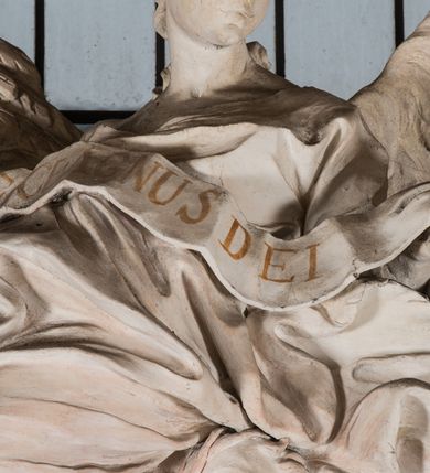 Zdjęcie nr 27: Na niskim postumencie, na rzucie prostokąta z łukiem nadwieszonym od przodu osadzony ołtarz prostopadłościenny. Antepedium marmoryzowane z owalnym polem i rytym przedstawieniem głowy św. Jana Chrzciciela na tacy spoczywającej na postumencie ze skrzyżowanymi szablą i krzyżem oraz banderolą z inskrypcją „ECCE AGNUS DEI”. Zasadniczą część ołtarza stanowi obraz w kształcie stojącego prostokąta w bogato profilowanej, szerokiej ramie, ujętej dwoma aniołami. Obaj stoją na skłębionych obłokach, ukazani frontalnie, w silnym kontrapoście. Ubrani są w przylegające do ich ciał szaty, modelowane swobodnie, silnie drapowane i marszczone. Anioł po lewej głowę zwraca w kierunku obrazu i unosi wysoko złożone w geście modlitwy ręce. Drugi anioł również zwraca się w kierunku obrazu, prawą rękę trzyma na piersi, lewą na wysokości bioder. Obaj mają rozłożyste skrzydła o gęstych piórach. Ramę wieńczy profilowany gzyms wyłamany półkoliście pośrodku z ukośnymi odcinkami na bokach o lekko wklęsłych krawędziach z podwieszonymi pod nie żłobkowanymi impostami, pod którymi unoszące je putta. Nad gzymsem pośrodku siedzi na obłoku z główkami aniołków anioł z rozłożystymi skrzydłami, trzymający w rękach banderolę z napisem „ECCE AGNUS DEI”. Po bokach gzymsu osadzone na nim obłoki z główkami aniołków. Ołtarz z wielobarwną marmoryzacją w dolnej części, czarną ramą i białą dekoracją rzeźbiarską.