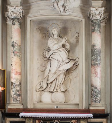 Zdjęcie nr 2: Na niewysokim stopniu prostopadłościenny ołtarz z antepedium, którego dekoracja wykonana w technice marmoryzacji (skaliola) w postaci owalnego pola z wyobrażeniem źródła życia wiecznego (dwa ptaki pijące wodę z fontanny) oraz pęków kwiatów. Nad ołtarzem nastawa z niemal pełnoplastyczną figurą Matki Boskiej Niepokalanej ujęta pilastrami i stojącymi przed nimi marmoryzowanymi kolumnami z kapitelami jońskimi podtrzymującymi szerokie belkowanie zwieńczone naczółkami. W środkowej części belkowania grupa rzeźbiarska z figurą Boga Ojca spoczywającego na obłokach trzymanych przez putta i anioły. Stojąca na obłokach i księżycu Matka Boska ukazana frontalnie, w kontrapoście, z rękami złożonymi do modlitwy. Jej twarz jest modelowana łagodnie, owalna, z długim nosem, otwartymi oczami. Nad głową wieniec z gwiazd. Ubrana jest w długą suknię i rozłożysty płaszcz o skłębionych połach, drapowanych plastycznie. Z tła wyłaniają się putta i uskrzydlone główki aniołów oraz promienie słońca. Postać Boga Ojca siedzącego na chmurach z rozwianymi szatami ukazana frontalnie. Prawą rękę wysuwa do przodu, w lewej trzyma berło. Ołtarz utrzymany w jasnej tonacji kolorystycznej, z elementami z czarnego marmuru i wielobarwnym antepedium w technice marmoryzacji. 