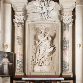 Zdjęcie nr 1: Na niewysokim stopniu prostopadłościenny ołtarz z antepedium, którego dekoracja wykonana w technice marmoryzacji (skaliola) w postaci owalnego pola z wyobrażeniem źródła życia wiecznego (dwa ptaki pijące wodę z fontanny) oraz pęków kwiatów. Nad ołtarzem nastawa z niemal pełnoplastyczną figurą Matki Boskiej Niepokalanej ujęta pilastrami i stojącymi przed nimi marmoryzowanymi kolumnami z kapitelami jońskimi podtrzymującymi szerokie belkowanie zwieńczone naczółkami. W środkowej części belkowania grupa rzeźbiarska z figurą Boga Ojca spoczywającego na obłokach trzymanych przez putta i anioły. Stojąca na obłokach i księżycu Matka Boska ukazana frontalnie, w kontrapoście, z rękami złożonymi do modlitwy. Jej twarz jest modelowana łagodnie, owalna, z długim nosem, otwartymi oczami. Nad głową wieniec z gwiazd. Ubrana jest w długą suknię i rozłożysty płaszcz o skłębionych połach, drapowanych plastycznie. Z tła wyłaniają się putta i uskrzydlone główki aniołów oraz promienie słońca. Postać Boga Ojca siedzącego na chmurach z rozwianymi szatami ukazana frontalnie. Prawą rękę wysuwa do przodu, w lewej trzyma berło. Ołtarz utrzymany w jasnej tonacji kolorystycznej, z elementami z czarnego marmuru i wielobarwnym antepedium w technice marmoryzacji. 