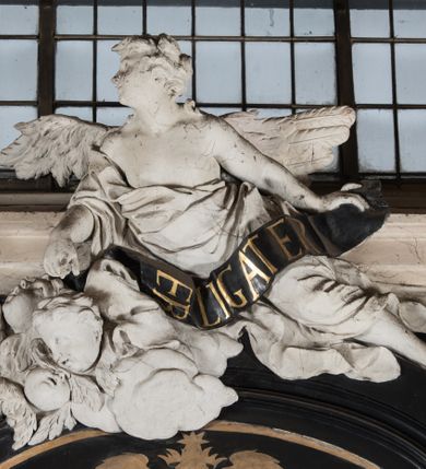 Zdjęcie nr 21: Na niskim postumencie, na rzucie prostokąta z łukiem nadwieszonym od przodu, prostopadłościenny ołtarz z antepedium ujętym szerokimi lizenami z romboidalnymi polami wypełnionymi drobnymi kwiatami, z owalnym polem pośrodku z podłużnymi palmami męczeństwa i bukietami kwiatów wokół. Nad ołtarzem nastawa z obrazem w kształcie stojącego prostokąta z przedstawieniem św. Piotra uwalnianego przez anioła z okowów. Ołtarz jest ujęty ukośnie dostawionymi postumentami z figurami dwóch aniołów flankujących obraz. Anioły ukazane w poruszonych pozach. Lewy stoi w silnym kontrapoście, z prawą nogą opartą na chmurze, z ciałem skierowanym ku środkowi osi ołtarza, z głową odwróconą w kierunku przeciwnym i wysuniętymi przed siebie rękami złożonymi do modlitwy. Jego twarz jest modelowana łagodnie, nos ma długi, usta rozwarte, a włosy skręcone, krótkie. Ubrany w obficie drapowany płaszcz, którego poły układają się w miękkie fałdy, dołem przechodzące między łydkami. Skrzydła ma rozłożyste, modelowane niesymetrycznie, gęsto upierzone. Drugi anioł klęczy na lewej nodze, wspartej na chmurze, zwrócony jest w kierunku obrazu, z prawą ręką przed sobą, a lewą odrzuconą do tyłu. Twarz ma pociągłą, nos wydatny, włosy krótkie, skręcone. Ubrany w długą suknię, drapowaną obficie, odsłaniającą lewe przedramię. Skrzydła rozłożyste, gęsto upierzone. Górna część ramy obrazu z ukośnie wyłamanym gzymsem z podwieszonymi pod nim impostami, podtrzymywanymi przez wyłaniające się z chmur anioły. Gzyms nad obrazem wyłamuje się półkoliście, na nim pośrodku leży anioł trzymający w lewej ręce banderolę ze złoconymi literami nietworzącymi logicznej całości, a prawą ręką wskazujący w kierunku wnętrza kaplicy. U jego boku skłębione chmury z trzema uskrzydlonymi główkami aniołków. Nad bocznymi partiami gzymsu siedzą uskrzydlone, nagie, pulchne aniołki, jeden z rękami złożonymi w geście modlitwy, drugi z rękami uniesionymi i rozłożonymi. Marmoryzacja utrzymana w kolorach brązów, rama i zwieńczenie czarne, dekoracja rzeźbiarska biała. 