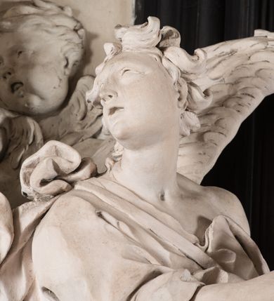 Zdjęcie nr 41: Na niskim postumencie, na rzucie prostokąta z łukiem nadwieszonym od przodu, prostopadłościenny ołtarz z antepedium ujętym szerokimi lizenami z romboidalnymi polami wypełnionymi drobnymi kwiatami, z owalnym polem pośrodku z podłużnymi palmami męczeństwa i bukietami kwiatów wokół. Nad ołtarzem nastawa z obrazem w kształcie stojącego prostokąta z przedstawieniem św. Piotra uwalnianego przez anioła z okowów. Ołtarz jest ujęty ukośnie dostawionymi postumentami z figurami dwóch aniołów flankujących obraz. Anioły ukazane w poruszonych pozach. Lewy stoi w silnym kontrapoście, z prawą nogą opartą na chmurze, z ciałem skierowanym ku środkowi osi ołtarza, z głową odwróconą w kierunku przeciwnym i wysuniętymi przed siebie rękami złożonymi do modlitwy. Jego twarz jest modelowana łagodnie, nos ma długi, usta rozwarte, a włosy skręcone, krótkie. Ubrany w obficie drapowany płaszcz, którego poły układają się w miękkie fałdy, dołem przechodzące między łydkami. Skrzydła ma rozłożyste, modelowane niesymetrycznie, gęsto upierzone. Drugi anioł klęczy na lewej nodze, wspartej na chmurze, zwrócony jest w kierunku obrazu, z prawą ręką przed sobą, a lewą odrzuconą do tyłu. Twarz ma pociągłą, nos wydatny, włosy krótkie, skręcone. Ubrany w długą suknię, drapowaną obficie, odsłaniającą lewe przedramię. Skrzydła rozłożyste, gęsto upierzone. Górna część ramy obrazu z ukośnie wyłamanym gzymsem z podwieszonymi pod nim impostami, podtrzymywanymi przez wyłaniające się z chmur anioły. Gzyms nad obrazem wyłamuje się półkoliście, na nim pośrodku leży anioł trzymający w lewej ręce banderolę ze złoconymi literami nietworzącymi logicznej całości, a prawą ręką wskazujący w kierunku wnętrza kaplicy. U jego boku skłębione chmury z trzema uskrzydlonymi główkami aniołków. Nad bocznymi partiami gzymsu siedzą uskrzydlone, nagie, pulchne aniołki, jeden z rękami złożonymi w geście modlitwy, drugi z rękami uniesionymi i rozłożonymi. Marmoryzacja utrzymana w kolorach brązów, rama i zwieńczenie czarne, dekoracja rzeźbiarska biała. 