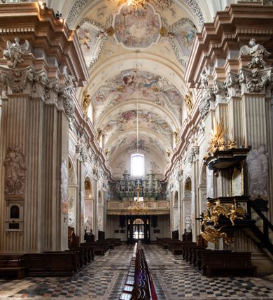 Zdjęcie nr 7: Kościół jest murowany z cegły, z użyciem ciosu kamiennego w partii cokołu i jako okładziny, tynkowany, z kryptami odpowiadającymi rzutowi. Założony jest na rzucie krzyża łacińskiego, z czteroprzęsłową nawą ujętą kaplicami, trójprzęsłowym prezbiterium zamknięty prosto, półtoraprzęsłowymi ramionami nawy poprzecznej, wysuniętymi poza korpus, zamknięte prosto i z kopułą na skrzyżowaniu naw. Przęsło fasady jest ujęte dwiema lokalnościami i dwiema wysuniętymi poza obręb korpusu wieżami z okrągłymi klatkami schodowymi z drewnianymi kręconymi schodami prowadzącymi do pomieszczeń nad lokalnościami. Do prezbiterium przylegają od południowego wschodu zakrystia, od północnego zachodu skarbiec, z składzikami na piętrze. 
Nawa jest artykułowa za pomocą pilastrów kompozytowych, zdwojonych w prezbiterium, transepcie i nawie głównej, zwielokrotniony w krzyżu kościoła, dźwigających wydatne belkowanie. Ściany kaplic są artykułowane za pomocą płytkich, opilastrowanych arkad; mury tarczowe kaplic wydzielone za pomocą gzymsu przełamującego się w narożnikach. Nawa i prezbiterium są sklepione kolebkami na gurtach z lunetami, transept sklepieniem kolebkowym na gurtach, kaplice pseudokopułami na pendentywach (w kaplicy Matki Boskiej i św. Katarzyny kopuły na rzucie elipsy z latarniami), zakrystia i skarbiec sklepieniem krzyżowo-kolebkowo, a przedsionki – nieckowym z lustrem. Okna nawy głównej i prezbiterium w profilowanych obramieniach, prostokątne, zamknięte łukiem segmentowym, okna kaplic są półkoliste. Bęben kopuły rozczłonkowany parami pilastrów kompozytowych, a między nimi rozlokowane naprzemian półkoliście zamknięte nisze i prostokątne okna. Do kościoła prowadzą drzwi wejściowe i para drzwi bocznych, z prezbiterium drzwi wejściowe do skarbca i zakrystii, z zakrystii na dziedziniec przy kościele. 
Fasada poprzedzona schodami, dwukondygnacyjna, dwuwieżowa, w dolnej kondygnacji siedmioosiowa, w górnej trójosiowa z dwiema wyodrębnionymi wieżami. Dolna kondygnacja rozczłonkowana zwojonymi pilastrami i parami kolumn flankujących oś środkową, o kapitelach kompozytowych, podtrzymujących wyłamujące się nad nimi belkowanie z wydatnym gzymsem i attyką, ponad belkowaniem w osi środkowej przerwany przyczółek. Górną kondygnację ujmują pary pilastrów ze stojącymi na ich tle wazonami, dźwigające wyłamujące się nad nimi belkowanie, w części środkowej zwieńczone przyczółkiem, w wieżach – miedzianymi hełmami. 
W części środkowej dolnej kondygnacji znajduje się zamknięta półkoliście wnęka z portalem, ujętym ukośnie dostawionymi pilastrami z konsolami podtrzymującymi naczółki i z tablicą z wklęsłymi bokami, zwieńczoną gzymsem w kształcie łuku nadwieszonego; ponad tablicą znajduje się półkoliste okno. Nad drzwiami bocznymi płaskorzeźbione postacie klęczących aniołów i kwadratowe okna, w wieżach wnęki z figurami św. Bernarda (po lewej) i św. Jana Kantego (po prawej). W części środkowej drugiej kondygnacji umieszczone jest prostokątne okno zamknięte łukiem segmentowym i zwieńczone przerwanym przyczółkiem. W polach bocznych nisze z figurami św. Kazimierza (po lewej) i św. Floriana (po prawej). Ściany boczne wzmocnione szkarpami, podzielone lizenami i pilastrami toskańskimi w dolnej kondygnacji prezbiterium oraz południowo wschodniej i północno zachodniej ścianie transeptu. Prezbiterium i ramiona transeptu zamknięte trójkątnymi szczytami z owalnymi oknami. Tambur kopuły rozczłonkowany lizenami, z oknami zamkniętymi łukami segmentowymi, w profilowanych obramieniach; ośmiopolową kopułę krytą blachą wieńczy latarnia opięta półkolumnami, pomiędzy którymi znajdują się wąskie okna zamknięte półkoliście. Nawę, prezbiterium i transept nakrywa dach siodłowy, nad pozostałymi pomieszczeniami dachy siodłowe, z wyjątkiem środkowych kaplic, w których kopułki wtopione zostały w dach i zwieńczone latarniami.  
