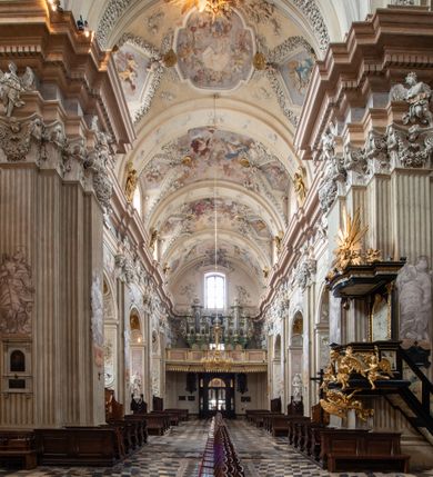Zdjęcie nr 9: Kościół jest murowany z cegły, z użyciem ciosu kamiennego w partii cokołu i jako okładziny, tynkowany, z kryptami odpowiadającymi rzutowi. Założony jest na rzucie krzyża łacińskiego, z czteroprzęsłową nawą ujętą kaplicami, trójprzęsłowym prezbiterium zamknięty prosto, półtoraprzęsłowymi ramionami nawy poprzecznej, wysuniętymi poza korpus, zamknięte prosto i z kopułą na skrzyżowaniu naw. Przęsło fasady jest ujęte dwiema lokalnościami i dwiema wysuniętymi poza obręb korpusu wieżami z okrągłymi klatkami schodowymi z drewnianymi kręconymi schodami prowadzącymi do pomieszczeń nad lokalnościami. Do prezbiterium przylegają od południowego wschodu zakrystia, od północnego zachodu skarbiec, z składzikami na piętrze. 
Nawa jest artykułowa za pomocą pilastrów kompozytowych, zdwojonych w prezbiterium, transepcie i nawie głównej, zwielokrotniony w krzyżu kościoła, dźwigających wydatne belkowanie. Ściany kaplic są artykułowane za pomocą płytkich, opilastrowanych arkad; mury tarczowe kaplic wydzielone za pomocą gzymsu przełamującego się w narożnikach. Nawa i prezbiterium są sklepione kolebkami na gurtach z lunetami, transept sklepieniem kolebkowym na gurtach, kaplice pseudokopułami na pendentywach (w kaplicy Matki Boskiej i św. Katarzyny kopuły na rzucie elipsy z latarniami), zakrystia i skarbiec sklepieniem krzyżowo-kolebkowo, a przedsionki – nieckowym z lustrem. Okna nawy głównej i prezbiterium w profilowanych obramieniach, prostokątne, zamknięte łukiem segmentowym, okna kaplic są półkoliste. Bęben kopuły rozczłonkowany parami pilastrów kompozytowych, a między nimi rozlokowane naprzemian półkoliście zamknięte nisze i prostokątne okna. Do kościoła prowadzą drzwi wejściowe i para drzwi bocznych, z prezbiterium drzwi wejściowe do skarbca i zakrystii, z zakrystii na dziedziniec przy kościele. 
Fasada poprzedzona schodami, dwukondygnacyjna, dwuwieżowa, w dolnej kondygnacji siedmioosiowa, w górnej trójosiowa z dwiema wyodrębnionymi wieżami. Dolna kondygnacja rozczłonkowana zwojonymi pilastrami i parami kolumn flankujących oś środkową, o kapitelach kompozytowych, podtrzymujących wyłamujące się nad nimi belkowanie z wydatnym gzymsem i attyką, ponad belkowaniem w osi środkowej przerwany przyczółek. Górną kondygnację ujmują pary pilastrów ze stojącymi na ich tle wazonami, dźwigające wyłamujące się nad nimi belkowanie, w części środkowej zwieńczone przyczółkiem, w wieżach – miedzianymi hełmami. 
W części środkowej dolnej kondygnacji znajduje się zamknięta półkoliście wnęka z portalem, ujętym ukośnie dostawionymi pilastrami z konsolami podtrzymującymi naczółki i z tablicą z wklęsłymi bokami, zwieńczoną gzymsem w kształcie łuku nadwieszonego; ponad tablicą znajduje się półkoliste okno. Nad drzwiami bocznymi płaskorzeźbione postacie klęczących aniołów i kwadratowe okna, w wieżach wnęki z figurami św. Bernarda (po lewej) i św. Jana Kantego (po prawej). W części środkowej drugiej kondygnacji umieszczone jest prostokątne okno zamknięte łukiem segmentowym i zwieńczone przerwanym przyczółkiem. W polach bocznych nisze z figurami św. Kazimierza (po lewej) i św. Floriana (po prawej). Ściany boczne wzmocnione szkarpami, podzielone lizenami i pilastrami toskańskimi w dolnej kondygnacji prezbiterium oraz południowo wschodniej i północno zachodniej ścianie transeptu. Prezbiterium i ramiona transeptu zamknięte trójkątnymi szczytami z owalnymi oknami. Tambur kopuły rozczłonkowany lizenami, z oknami zamkniętymi łukami segmentowymi, w profilowanych obramieniach; ośmiopolową kopułę krytą blachą wieńczy latarnia opięta półkolumnami, pomiędzy którymi znajdują się wąskie okna zamknięte półkoliście. Nawę, prezbiterium i transept nakrywa dach siodłowy, nad pozostałymi pomieszczeniami dachy siodłowe, z wyjątkiem środkowych kaplic, w których kopułki wtopione zostały w dach i zwieńczone latarniami.  
