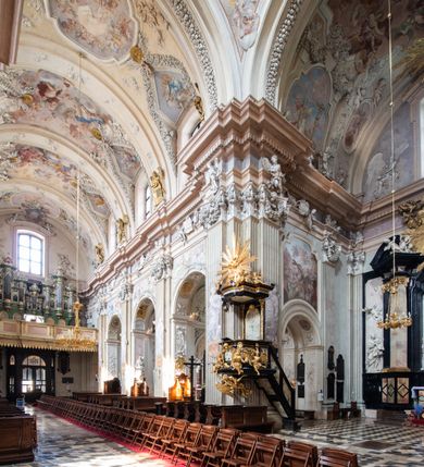 Zdjęcie nr 17: Kościół jest murowany z cegły, z użyciem ciosu kamiennego w partii cokołu i jako okładziny, tynkowany, z kryptami odpowiadającymi rzutowi. Założony jest na rzucie krzyża łacińskiego, z czteroprzęsłową nawą ujętą kaplicami, trójprzęsłowym prezbiterium zamknięty prosto, półtoraprzęsłowymi ramionami nawy poprzecznej, wysuniętymi poza korpus, zamknięte prosto i z kopułą na skrzyżowaniu naw. Przęsło fasady jest ujęte dwiema lokalnościami i dwiema wysuniętymi poza obręb korpusu wieżami z okrągłymi klatkami schodowymi z drewnianymi kręconymi schodami prowadzącymi do pomieszczeń nad lokalnościami. Do prezbiterium przylegają od południowego wschodu zakrystia, od północnego zachodu skarbiec, z składzikami na piętrze. 
Nawa jest artykułowa za pomocą pilastrów kompozytowych, zdwojonych w prezbiterium, transepcie i nawie głównej, zwielokrotniony w krzyżu kościoła, dźwigających wydatne belkowanie. Ściany kaplic są artykułowane za pomocą płytkich, opilastrowanych arkad; mury tarczowe kaplic wydzielone za pomocą gzymsu przełamującego się w narożnikach. Nawa i prezbiterium są sklepione kolebkami na gurtach z lunetami, transept sklepieniem kolebkowym na gurtach, kaplice pseudokopułami na pendentywach (w kaplicy Matki Boskiej i św. Katarzyny kopuły na rzucie elipsy z latarniami), zakrystia i skarbiec sklepieniem krzyżowo-kolebkowo, a przedsionki – nieckowym z lustrem. Okna nawy głównej i prezbiterium w profilowanych obramieniach, prostokątne, zamknięte łukiem segmentowym, okna kaplic są półkoliste. Bęben kopuły rozczłonkowany parami pilastrów kompozytowych, a między nimi rozlokowane naprzemian półkoliście zamknięte nisze i prostokątne okna. Do kościoła prowadzą drzwi wejściowe i para drzwi bocznych, z prezbiterium drzwi wejściowe do skarbca i zakrystii, z zakrystii na dziedziniec przy kościele. 
Fasada poprzedzona schodami, dwukondygnacyjna, dwuwieżowa, w dolnej kondygnacji siedmioosiowa, w górnej trójosiowa z dwiema wyodrębnionymi wieżami. Dolna kondygnacja rozczłonkowana zwojonymi pilastrami i parami kolumn flankujących oś środkową, o kapitelach kompozytowych, podtrzymujących wyłamujące się nad nimi belkowanie z wydatnym gzymsem i attyką, ponad belkowaniem w osi środkowej przerwany przyczółek. Górną kondygnację ujmują pary pilastrów ze stojącymi na ich tle wazonami, dźwigające wyłamujące się nad nimi belkowanie, w części środkowej zwieńczone przyczółkiem, w wieżach – miedzianymi hełmami. 
W części środkowej dolnej kondygnacji znajduje się zamknięta półkoliście wnęka z portalem, ujętym ukośnie dostawionymi pilastrami z konsolami podtrzymującymi naczółki i z tablicą z wklęsłymi bokami, zwieńczoną gzymsem w kształcie łuku nadwieszonego; ponad tablicą znajduje się półkoliste okno. Nad drzwiami bocznymi płaskorzeźbione postacie klęczących aniołów i kwadratowe okna, w wieżach wnęki z figurami św. Bernarda (po lewej) i św. Jana Kantego (po prawej). W części środkowej drugiej kondygnacji umieszczone jest prostokątne okno zamknięte łukiem segmentowym i zwieńczone przerwanym przyczółkiem. W polach bocznych nisze z figurami św. Kazimierza (po lewej) i św. Floriana (po prawej). Ściany boczne wzmocnione szkarpami, podzielone lizenami i pilastrami toskańskimi w dolnej kondygnacji prezbiterium oraz południowo wschodniej i północno zachodniej ścianie transeptu. Prezbiterium i ramiona transeptu zamknięte trójkątnymi szczytami z owalnymi oknami. Tambur kopuły rozczłonkowany lizenami, z oknami zamkniętymi łukami segmentowymi, w profilowanych obramieniach; ośmiopolową kopułę krytą blachą wieńczy latarnia opięta półkolumnami, pomiędzy którymi znajdują się wąskie okna zamknięte półkoliście. Nawę, prezbiterium i transept nakrywa dach siodłowy, nad pozostałymi pomieszczeniami dachy siodłowe, z wyjątkiem środkowych kaplic, w których kopułki wtopione zostały w dach i zwieńczone latarniami.  
