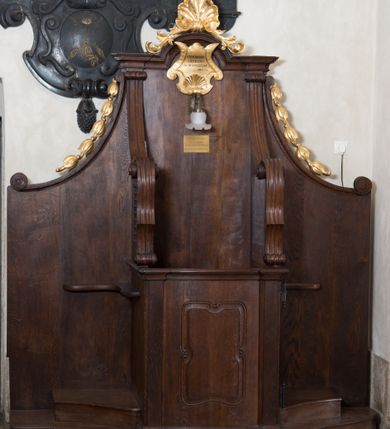 Zdjęcie nr 5: Para konfesjonałów o lekko wybrzuszonym przodzie i wklęsłych bokach, z wysokim zapleckiem ze ściętymi, wklęsłymi bokami, ujętymi spływami wolutowymi. Siedzisko z drzwiczkami ozdobionymi wklęsłą ramą z łukowatymi wcięciami na każdym boku i bokami wyciętymi linią wklęsło-wypukłą, z kratkami. Środkowa część zaplecka zwieńczona profilowanym gzymsem z łukiem nadwieszonymi, w którym znajduje się złocony kartusz ujęty karbowaną taśmą i muszlą palmetową z inskrypcją „KTÓRYCH ODPUŚCICIE / GRZECHY / SĄ IM ODPUSZCZONE / JAN XX. 23”. Nad łukiem muszla palmetowa ujęta wiciami akantu. Wzdłuż spływów wolutowych po bokach złocone kampanule. Do zaplecka, nad siedziskiem zamontowana lampka elektryczna i tabliczka z inskrypcją „W TYM KONFESJONALE SPOWIADAŁ / bł(ogosławiony) Jan Paweł II / ks(iądz) Kardynał Karol Wojtyła / gdy był Pasterzem Kościoła Krakowskiego.”
