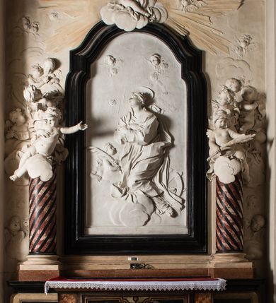 Zdjęcie nr 2: Na trójstopniowej podstawie osadzona prostopadłościenna mensa ołtarza ujęta lekko wybrzuszającymi się postumentami. Antepedium wykonane w technice marmoryzacji (skaliola) z owalnym polem pośrodku, z wyobrażeniem leżącego nagiego Dzieciątka Jezus z krzyżykiem w lewej dłoni i pierścieniem z krzyżem w prawej, pozostałe naroża wypełnione bukietami kwiatów. Na osi mensy nastawa w formie profilowanej ramy prostokątnej zamkniętej łukiem wklęsło-wypukłym, nadwieszonym, wypełnionej płaskorzeźbą z postacią św. Katarzyny. Święta ukazana w trzech czwartych, w pozycji klęczącej, na chmurach, z rękami złożonymi na piersi, z głową lekko odchyloną w głąb kompozycji. Jej twarz jest owalna, o pełnych policzkach, ze smukłym nosem, głębokimi oczodołami i otwartymi ustami. Na głowie włosy modelowane sumarycznie, długie, opadające na plecy oraz korona otwarta. Święta jest ubrana w długą suknię i obficie fałdowany płaszcz. Za jej plecami widoczny fragment koła – atrybutu jej męczeństwa, z przodu wyłaniające się płaskorzeźbione putta, z których jedno trzyma gałąź palmy. W górnej części kompozycji modelowane w płytkim reliefie chmury z widocznymi między nimi promieniami oraz uskrzydlone główki aniołków. Po bokach kolumny osadzone na bazach, ścięte w połowie wysokości ramy, zakończone skłębionymi obłokami chmur i siedzącymi na nich aniołkami. Lewy ukazany w pozycji półleżącej, z prawą ręką na piersi, a lewą wyciągniętą w kierunku osi ołtarza, z głową skierowaną w jego prawą stronę. Anioł po przeciwnej stronie siedzi na obłoku okrakiem i trzyma w ręku palmę męczeństwa. W tle obu aniołów płaskorzeźbione grupy puttów z banderolami z nieczytelnymi inskrypcjami. Nastawę wieńczy gloria promienista, której promienie wchodzą w partię okna, z siedzącą na obłokach postacią małego Jezusa z kulą ziemską przy boku i z prawą ręką uniesioną w geście błogosławieństwa, ubranego w rozwiany na plecach płaszcz. Kompozycję uzupełniają płaskorzeźbione grupy uskrzydlonych aniołków. Ołtarz z elementami czarnego i białego marmuru, barwnej marmoryzacji i białego, częściowo złoconego stiuku. 