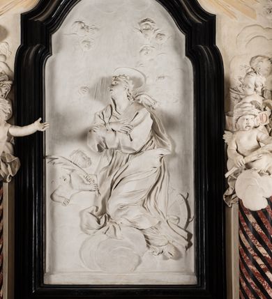 Zdjęcie nr 14: Na trójstopniowej podstawie osadzona prostopadłościenna mensa ołtarza ujęta lekko wybrzuszającymi się postumentami. Antepedium wykonane w technice marmoryzacji (skaliola) z owalnym polem pośrodku, z wyobrażeniem leżącego nagiego Dzieciątka Jezus z krzyżykiem w lewej dłoni i pierścieniem z krzyżem w prawej, pozostałe naroża wypełnione bukietami kwiatów. Na osi mensy nastawa w formie profilowanej ramy prostokątnej zamkniętej łukiem wklęsło-wypukłym, nadwieszonym, wypełnionej płaskorzeźbą z postacią św. Katarzyny. Święta ukazana w trzech czwartych, w pozycji klęczącej, na chmurach, z rękami złożonymi na piersi, z głową lekko odchyloną w głąb kompozycji. Jej twarz jest owalna, o pełnych policzkach, ze smukłym nosem, głębokimi oczodołami i otwartymi ustami. Na głowie włosy modelowane sumarycznie, długie, opadające na plecy oraz korona otwarta. Święta jest ubrana w długą suknię i obficie fałdowany płaszcz. Za jej plecami widoczny fragment koła – atrybutu jej męczeństwa, z przodu wyłaniające się płaskorzeźbione putta, z których jedno trzyma gałąź palmy. W górnej części kompozycji modelowane w płytkim reliefie chmury z widocznymi między nimi promieniami oraz uskrzydlone główki aniołków. Po bokach kolumny osadzone na bazach, ścięte w połowie wysokości ramy, zakończone skłębionymi obłokami chmur i siedzącymi na nich aniołkami. Lewy ukazany w pozycji półleżącej, z prawą ręką na piersi, a lewą wyciągniętą w kierunku osi ołtarza, z głową skierowaną w jego prawą stronę. Anioł po przeciwnej stronie siedzi na obłoku okrakiem i trzyma w ręku palmę męczeństwa. W tle obu aniołów płaskorzeźbione grupy puttów z banderolami z nieczytelnymi inskrypcjami. Nastawę wieńczy gloria promienista, której promienie wchodzą w partię okna, z siedzącą na obłokach postacią małego Jezusa z kulą ziemską przy boku i z prawą ręką uniesioną w geście błogosławieństwa, ubranego w rozwiany na plecach płaszcz. Kompozycję uzupełniają płaskorzeźbione grupy uskrzydlonych aniołków. Ołtarz z elementami czarnego i białego marmuru, barwnej marmoryzacji i białego, częściowo złoconego stiuku. 