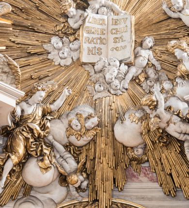 Zdjęcie nr 23: Zasadniczy ołtarz jest prostopadłościenny, z antepedium w technice skalioli, z owalnym polem, w którym znajduje się medalion obwiedziony kwiatami. Wydzielone wokół niego geometryczne pola pokryte są białą i czerwoną marmoryzacją. Za ołtarzem znajduje się monumentalna nastawa, składająca się z podwójnych cokołów, dwóch tylnych, ustawionych równolegle do prezbiterium, dwóch przednich ustawionych ukośnie, na których stoją osadzone na tle pilastrów kolumny o spiralnych trzonach. Po bokach kolumn unoszące się na chmurach uskrzydlone anioły w miękko modelowanych szatach. Trzony kolumn ozdobione wijącą się wokół nich wicią roślinną, ich kapitele w porządku kompozytowym podtrzymują belkowanie. Nad zewnętrznymi kolumnami unoszą się faliste naczółki, zaś nad wewnętrznymi siedzące anioły. Nad ołtarzem posadowiono dużych rozmiarów glorię promienistą z grupą aniołów i puttów. Pośrodku otwarta księga trzymana przez anioły z napisem: LIBER GENERATIONIS IESV CHRISTI. W polu środkowym nisza na obraz w kształcie prostokąta zamkniętego półkoliście, obwiedzionego rzeźbioną ramą z motywem liści lauru; na obrazie przedstawiona św. Anna Samotrzeć. Ołtarz flankują wsunięte do przodu figury św. Wojciecha i św. Stanisława, stojące na dostawionych do ścian bocznych prezbiterium cokołach, wybrzuszających się w górnej części. 