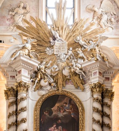 Zdjęcie nr 13: Zasadniczy ołtarz jest prostopadłościenny, z antepedium w technice skalioli, z owalnym polem, w którym znajduje się medalion obwiedziony kwiatami. Wydzielone wokół niego geometryczne pola pokryte są białą i czerwoną marmoryzacją. Za ołtarzem znajduje się monumentalna nastawa, składająca się z podwójnych cokołów, dwóch tylnych, ustawionych równolegle do prezbiterium, dwóch przednich ustawionych ukośnie, na których stoją osadzone na tle pilastrów kolumny o spiralnych trzonach. Po bokach kolumn unoszące się na chmurach uskrzydlone anioły w miękko modelowanych szatach. Trzony kolumn ozdobione wijącą się wokół nich wicią roślinną, ich kapitele w porządku kompozytowym podtrzymują belkowanie. Nad zewnętrznymi kolumnami unoszą się faliste naczółki, zaś nad wewnętrznymi siedzące anioły. Nad ołtarzem posadowiono dużych rozmiarów glorię promienistą z grupą aniołów i puttów. Pośrodku otwarta księga trzymana przez anioły z napisem: LIBER GENERATIONIS IESV CHRISTI. W polu środkowym nisza na obraz w kształcie prostokąta zamkniętego półkoliście, obwiedzionego rzeźbioną ramą z motywem liści lauru; na obrazie przedstawiona św. Anna Samotrzeć. Ołtarz flankują wsunięte do przodu figury św. Wojciecha i św. Stanisława, stojące na dostawionych do ścian bocznych prezbiterium cokołach, wybrzuszających się w górnej części. 