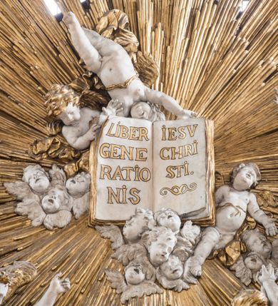 Zdjęcie nr 25: Zasadniczy ołtarz jest prostopadłościenny, z antepedium w technice skalioli, z owalnym polem, w którym znajduje się medalion obwiedziony kwiatami. Wydzielone wokół niego geometryczne pola pokryte są białą i czerwoną marmoryzacją. Za ołtarzem znajduje się monumentalna nastawa, składająca się z podwójnych cokołów, dwóch tylnych, ustawionych równolegle do prezbiterium, dwóch przednich ustawionych ukośnie, na których stoją osadzone na tle pilastrów kolumny o spiralnych trzonach. Po bokach kolumn unoszące się na chmurach uskrzydlone anioły w miękko modelowanych szatach. Trzony kolumn ozdobione wijącą się wokół nich wicią roślinną, ich kapitele w porządku kompozytowym podtrzymują belkowanie. Nad zewnętrznymi kolumnami unoszą się faliste naczółki, zaś nad wewnętrznymi siedzące anioły. Nad ołtarzem posadowiono dużych rozmiarów glorię promienistą z grupą aniołów i puttów. Pośrodku otwarta księga trzymana przez anioły z napisem: LIBER GENERATIONIS IESV CHRISTI. W polu środkowym nisza na obraz w kształcie prostokąta zamkniętego półkoliście, obwiedzionego rzeźbioną ramą z motywem liści lauru; na obrazie przedstawiona św. Anna Samotrzeć. Ołtarz flankują wsunięte do przodu figury św. Wojciecha i św. Stanisława, stojące na dostawionych do ścian bocznych prezbiterium cokołach, wybrzuszających się w górnej części. 