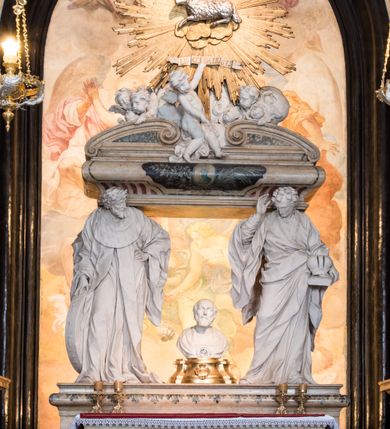 Zdjęcie nr 19: Na prostopadłościennej, wysokiej podstawie na kilkustopniowym podwyższeniu z dostawionym do niej ołtarzem, stoją personifikacje czterech Wydziałów Uniwersytetu – Teologii, Prawa, Medycyny i Sztuk Wyzwolonych, podtrzymujące trumnę z figurą putta w otoczeniu obłoków, które prawą ręką wskazuje na świętego, a lewą na posrebrzaną rzeźbę Baranka Apokaliptycznego na tle glorii i znajdującą poniżej banderolę z napisem: "Et lucerna eius est Agnus". Trumna ma kształt wybrzuszonego dołem sarkofagu z wiekiem ujętym dwiema wolutami. Ołtarz z antepedium wykonanym w technice skalioli z wyobrażeniem uskrzydlonego anioła trzymającego w rękach wieńce z kwiatów i lauru otoczonego witkami palm i rozlokowany i swobodnie pękami różnokolorowych kwiatków. Wokół grupy ustawiono cztery wolnostojące wysokie kolumny posadowione na wysokich postumentach, których ścianki są ozdobione czarnymi medalionami z motywami kwiatów wykonanymi w technice skalioli. Kolumny mają  skręcone trzony i kompozytowe kapitele, dźwigające odcinki belkowania, na których umieszczono figury czterech świętych Janów – Jana Chrzciciela, Jana Ewangelisty, Jana Kantego i Jana z Damaszku. 



