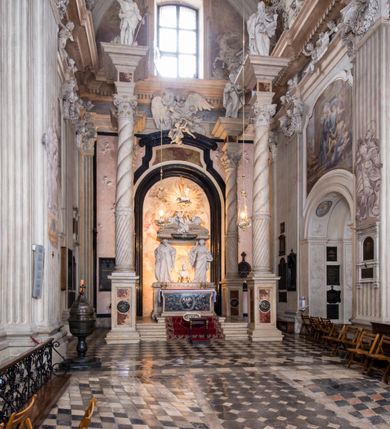 Zdjęcie nr 13: Na prostopadłościennej, wysokiej podstawie na kilkustopniowym podwyższeniu z dostawionym do niej ołtarzem, stoją personifikacje czterech Wydziałów Uniwersytetu – Teologii, Prawa, Medycyny i Sztuk Wyzwolonych, podtrzymujące trumnę z figurą putta w otoczeniu obłoków, które prawą ręką wskazuje na świętego, a lewą na posrebrzaną rzeźbę Baranka Apokaliptycznego na tle glorii i znajdującą poniżej banderolę z napisem: &quot;Et lucerna eius est Agnus&quot;. Trumna ma kształt wybrzuszonego dołem sarkofagu z wiekiem ujętym dwiema wolutami. Ołtarz z antepedium wykonanym w technice skalioli z wyobrażeniem uskrzydlonego anioła trzymającego w rękach wieńce z kwiatów i lauru otoczonego witkami palm i rozlokowany i swobodnie pękami różnokolorowych kwiatków. Wokół grupy ustawiono cztery wolnostojące wysokie kolumny posadowione na wysokich postumentach, których ścianki są ozdobione czarnymi medalionami z motywami kwiatów wykonanymi w technice skalioli. Kolumny mają  skręcone trzony i kompozytowe kapitele, dźwigające odcinki belkowania, na których umieszczono figury czterech świętych Janów – Jana Chrzciciela, Jana Ewangelisty, Jana Kantego i Jana z Damaszku. 



