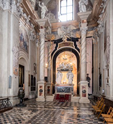 Zdjęcie nr 17: Na prostopadłościennej, wysokiej podstawie na kilkustopniowym podwyższeniu z dostawionym do niej ołtarzem, stoją personifikacje czterech Wydziałów Uniwersytetu – Teologii, Prawa, Medycyny i Sztuk Wyzwolonych, podtrzymujące trumnę z figurą putta w otoczeniu obłoków, które prawą ręką wskazuje na świętego, a lewą na posrebrzaną rzeźbę Baranka Apokaliptycznego na tle glorii i znajdującą poniżej banderolę z napisem: &quot;Et lucerna eius est Agnus&quot;. Trumna ma kształt wybrzuszonego dołem sarkofagu z wiekiem ujętym dwiema wolutami. Ołtarz z antepedium wykonanym w technice skalioli z wyobrażeniem uskrzydlonego anioła trzymającego w rękach wieńce z kwiatów i lauru otoczonego witkami palm i rozlokowany i swobodnie pękami różnokolorowych kwiatków. Wokół grupy ustawiono cztery wolnostojące wysokie kolumny posadowione na wysokich postumentach, których ścianki są ozdobione czarnymi medalionami z motywami kwiatów wykonanymi w technice skalioli. Kolumny mają  skręcone trzony i kompozytowe kapitele, dźwigające odcinki belkowania, na których umieszczono figury czterech świętych Janów – Jana Chrzciciela, Jana Ewangelisty, Jana Kantego i Jana z Damaszku. 



