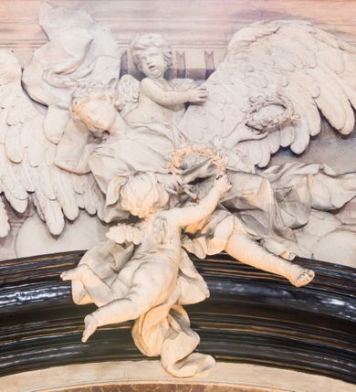 Zdjęcie nr 14: Na prostopadłościennej, wysokiej podstawie na kilkustopniowym podwyższeniu z dostawionym do niej ołtarzem, stoją personifikacje czterech Wydziałów Uniwersytetu – Teologii, Prawa, Medycyny i Sztuk Wyzwolonych, podtrzymujące trumnę z figurą putta w otoczeniu obłoków, które prawą ręką wskazuje na świętego, a lewą na posrebrzaną rzeźbę Baranka Apokaliptycznego na tle glorii i znajdującą poniżej banderolę z napisem: &quot;Et lucerna eius est Agnus&quot;. Trumna ma kształt wybrzuszonego dołem sarkofagu z wiekiem ujętym dwiema wolutami. Ołtarz z antepedium wykonanym w technice skalioli z wyobrażeniem uskrzydlonego anioła trzymającego w rękach wieńce z kwiatów i lauru otoczonego witkami palm i rozlokowany i swobodnie pękami różnokolorowych kwiatków. Wokół grupy ustawiono cztery wolnostojące wysokie kolumny posadowione na wysokich postumentach, których ścianki są ozdobione czarnymi medalionami z motywami kwiatów wykonanymi w technice skalioli. Kolumny mają  skręcone trzony i kompozytowe kapitele, dźwigające odcinki belkowania, na których umieszczono figury czterech świętych Janów – Jana Chrzciciela, Jana Ewangelisty, Jana Kantego i Jana z Damaszku. 



