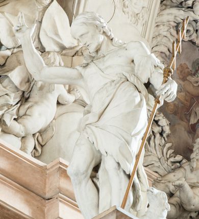 Zdjęcie nr 6: Na prostopadłościennej, wysokiej podstawie na kilkustopniowym podwyższeniu z dostawionym do niej ołtarzem, stoją personifikacje czterech Wydziałów Uniwersytetu – Teologii, Prawa, Medycyny i Sztuk Wyzwolonych, podtrzymujące trumnę z figurą putta w otoczeniu obłoków, które prawą ręką wskazuje na świętego, a lewą na posrebrzaną rzeźbę Baranka Apokaliptycznego na tle glorii i znajdującą poniżej banderolę z napisem: &quot;Et lucerna eius est Agnus&quot;. Trumna ma kształt wybrzuszonego dołem sarkofagu z wiekiem ujętym dwiema wolutami. Ołtarz z antepedium wykonanym w technice skalioli z wyobrażeniem uskrzydlonego anioła trzymającego w rękach wieńce z kwiatów i lauru otoczonego witkami palm i rozlokowany i swobodnie pękami różnokolorowych kwiatków. Wokół grupy ustawiono cztery wolnostojące wysokie kolumny posadowione na wysokich postumentach, których ścianki są ozdobione czarnymi medalionami z motywami kwiatów wykonanymi w technice skalioli. Kolumny mają  skręcone trzony i kompozytowe kapitele, dźwigające odcinki belkowania, na których umieszczono figury czterech świętych Janów – Jana Chrzciciela, Jana Ewangelisty, Jana Kantego i Jana z Damaszku. 



