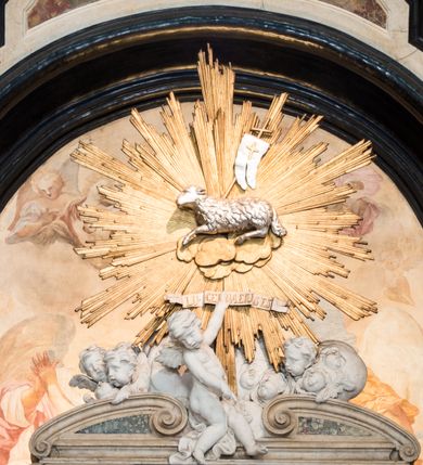 Zdjęcie nr 21: Na prostopadłościennej, wysokiej podstawie na kilkustopniowym podwyższeniu z dostawionym do niej ołtarzem, stoją personifikacje czterech Wydziałów Uniwersytetu – Teologii, Prawa, Medycyny i Sztuk Wyzwolonych, podtrzymujące trumnę z figurą putta w otoczeniu obłoków, które prawą ręką wskazuje na świętego, a lewą na posrebrzaną rzeźbę Baranka Apokaliptycznego na tle glorii i znajdującą poniżej banderolę z napisem: "Et lucerna eius est Agnus". Trumna ma kształt wybrzuszonego dołem sarkofagu z wiekiem ujętym dwiema wolutami. Ołtarz z antepedium wykonanym w technice skalioli z wyobrażeniem uskrzydlonego anioła trzymającego w rękach wieńce z kwiatów i lauru otoczonego witkami palm i rozlokowany i swobodnie pękami różnokolorowych kwiatków. Wokół grupy ustawiono cztery wolnostojące wysokie kolumny posadowione na wysokich postumentach, których ścianki są ozdobione czarnymi medalionami z motywami kwiatów wykonanymi w technice skalioli. Kolumny mają  skręcone trzony i kompozytowe kapitele, dźwigające odcinki belkowania, na których umieszczono figury czterech świętych Janów – Jana Chrzciciela, Jana Ewangelisty, Jana Kantego i Jana z Damaszku. 



