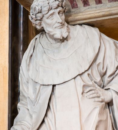 Zdjęcie nr 10: Na prostopadłościennej, wysokiej podstawie na kilkustopniowym podwyższeniu z dostawionym do niej ołtarzem, stoją personifikacje czterech Wydziałów Uniwersytetu – Teologii, Prawa, Medycyny i Sztuk Wyzwolonych, podtrzymujące trumnę z figurą putta w otoczeniu obłoków, które prawą ręką wskazuje na świętego, a lewą na posrebrzaną rzeźbę Baranka Apokaliptycznego na tle glorii i znajdującą poniżej banderolę z napisem: &quot;Et lucerna eius est Agnus&quot;. Trumna ma kształt wybrzuszonego dołem sarkofagu z wiekiem ujętym dwiema wolutami. Ołtarz z antepedium wykonanym w technice skalioli z wyobrażeniem uskrzydlonego anioła trzymającego w rękach wieńce z kwiatów i lauru otoczonego witkami palm i rozlokowany i swobodnie pękami różnokolorowych kwiatków. Wokół grupy ustawiono cztery wolnostojące wysokie kolumny posadowione na wysokich postumentach, których ścianki są ozdobione czarnymi medalionami z motywami kwiatów wykonanymi w technice skalioli. Kolumny mają  skręcone trzony i kompozytowe kapitele, dźwigające odcinki belkowania, na których umieszczono figury czterech świętych Janów – Jana Chrzciciela, Jana Ewangelisty, Jana Kantego i Jana z Damaszku. 



