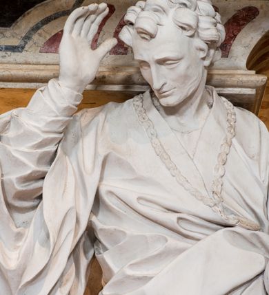 Zdjęcie nr 12: Na prostopadłościennej, wysokiej podstawie na kilkustopniowym podwyższeniu z dostawionym do niej ołtarzem, stoją personifikacje czterech Wydziałów Uniwersytetu – Teologii, Prawa, Medycyny i Sztuk Wyzwolonych, podtrzymujące trumnę z figurą putta w otoczeniu obłoków, które prawą ręką wskazuje na świętego, a lewą na posrebrzaną rzeźbę Baranka Apokaliptycznego na tle glorii i znajdującą poniżej banderolę z napisem: &quot;Et lucerna eius est Agnus&quot;. Trumna ma kształt wybrzuszonego dołem sarkofagu z wiekiem ujętym dwiema wolutami. Ołtarz z antepedium wykonanym w technice skalioli z wyobrażeniem uskrzydlonego anioła trzymającego w rękach wieńce z kwiatów i lauru otoczonego witkami palm i rozlokowany i swobodnie pękami różnokolorowych kwiatków. Wokół grupy ustawiono cztery wolnostojące wysokie kolumny posadowione na wysokich postumentach, których ścianki są ozdobione czarnymi medalionami z motywami kwiatów wykonanymi w technice skalioli. Kolumny mają  skręcone trzony i kompozytowe kapitele, dźwigające odcinki belkowania, na których umieszczono figury czterech świętych Janów – Jana Chrzciciela, Jana Ewangelisty, Jana Kantego i Jana z Damaszku. 



