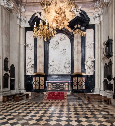 Zdjęcie nr 2: Ołtarz ustawiony na trójstopniowym podeście, prostopadłościenny, z antepedium z dekoracją w technice skaliola z pól geometrycznych dzielonych listwami i wypełniony drobnymi, wielobarwnymi kwiatkami. Za ołtarzem monumentalna nastawa utworzona z pilastrów i ukośnie ustawionych filarów stojących na wysokich postumentach. Ich trzony są kanelowane, a kapitele utworzone z jońskich wolut i przewiązanych taśmą lilijek. Nad kapitelami wznosi się belkowanie, nad zewnętrznymi pilastrami zwieńczone falistymi naczółkami, nad wewnętrznymi grupami uskrzydlonych puttów. Pośrodku znajduje się prostokątna wnęka zakończona łukiem półkolistym, nadwieszonym, w profilowanej ramie. W niej stiukowy relief przedstawiający Chwałę Krzyża Świętego adorowanego przez Matkę Boską z martwym Chrystusem spoczywającym na jej kolanach oraz przez św. Jana Ewangelistę odwróconego do widza plecami. Maria ukazana frontalnie, ubrana w rozłożystą suknię i  płaszcz zarzucony na głowę, z prawą ręką dotykająca głowy Jezusa, z lewą wyciągniętą do przodu. Jan przedstawiony w wykroku, z rękami wysuniętymi przed siebie, ubrany w suknię i płaszcz odkrywający lewy bark. Za Chrystusem ukazany anioł trzymający jego rękę. Nad nimi grupa aniołów unoszących krzyż. Ponad wnęką widnieje promienista, złocona gloria, z postacią Boga Ojca zasiadającego w chmurach, z gołębicą Ducha Świętego, puttami i główkami aniołów. Po bokach stoją figury św. Weroniki i św. Longina. Święta jest ubrana w długą, ekspresyjnie marszczoną suknię, w rękach trzyma rozłożoną chustę z twarzą Chrystusa. Longin jest przestawiony w zbroi rzymskiego legionisty, z długim, efektownie formowanym płaszczem, z lanca w prawej dłoni. 
