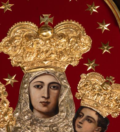 Zdjęcie nr 5: Obraz Matki Boskiej z Dzieciątkiem w kształcie stojącego prostokąta zamkniętego łukiem nadwieszonym. W centrum przedstawiona została Maria, trzymająca na lewej ręce Dzieciątko Jezus, z berłem w prawej. Maria spogląda na widza, ma owalną twarz o dużych, brązowych oczach, wąskim nosie i niewielkich, kształtnych ustach. Dzieciątko patrzy przed siebie, prawą ręką czyni gest błogosławieństwa, lewą trzyma rękę Marii. Poza widocznymi twarzami, rękami oraz stopami Dzieciątka resztę postaci zakrywają srebrne sukienki i złocone korony. Postacie zostały umieszczone na tle wykonanym z czerwonej tkaniny. Sukienki pokrywa gęsty wzór liściasto-kwiatowy złożony z rozwiniętych kwiatów i bujnych, mięsistych liści, z szerokim lamowaniem zdobionym rombowymi kaboszonami i krzyżykami. Dolna partia jest nowsza, dosztukowana i pokryta podobnym, choć rzadszym wzorem. Korony są złocone, wysokie, wypukłe, częściowo puklowane i trybowane w ornament z bujnej wici akantowej, nad czołem zdobione szlakiem z kaboszonów, a u  góry perełkowaniem i zwieńczone kulą z krzyżem. Wokół głowy Marii dodatkowo 12 gwiazdek. Matka Boska w prawej dłoni trzyma berło o rękojeści zdobionej dołem liśćmi akantu, a u góry rombowymi kaboszonami. 