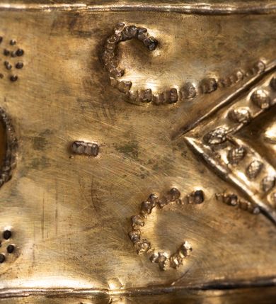 Zdjęcie nr 6: Obraz Matki Boskiej z Dzieciątkiem w kształcie stojącego prostokąta zamkniętego łukiem nadwieszonym. W centrum przedstawiona została Maria, trzymająca na lewej ręce Dzieciątko Jezus, z berłem w prawej. Maria spogląda na widza, ma owalną twarz o dużych, brązowych oczach, wąskim nosie i niewielkich, kształtnych ustach. Dzieciątko patrzy przed siebie, prawą ręką czyni gest błogosławieństwa, lewą trzyma rękę Marii. Poza widocznymi twarzami, rękami oraz stopami Dzieciątka resztę postaci zakrywają srebrne sukienki i złocone korony. Postacie zostały umieszczone na tle wykonanym z czerwonej tkaniny. Sukienki pokrywa gęsty wzór liściasto-kwiatowy złożony z rozwiniętych kwiatów i bujnych, mięsistych liści, z szerokim lamowaniem zdobionym rombowymi kaboszonami i krzyżykami. Dolna partia jest nowsza, dosztukowana i pokryta podobnym, choć rzadszym wzorem. Korony są złocone, wysokie, wypukłe, częściowo puklowane i trybowane w ornament z bujnej wici akantowej, nad czołem zdobione szlakiem z kaboszonów, a u  góry perełkowaniem i zwieńczone kulą z krzyżem. Wokół głowy Marii dodatkowo 12 gwiazdek. Matka Boska w prawej dłoni trzyma berło o rękojeści zdobionej dołem liśćmi akantu, a u góry rombowymi kaboszonami. 