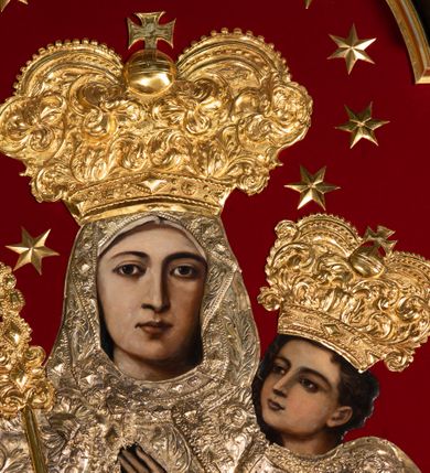 Zdjęcie nr 4: Obraz Matki Boskiej z Dzieciątkiem w kształcie stojącego prostokąta zamkniętego łukiem nadwieszonym. W centrum przedstawiona została Maria, trzymająca na lewej ręce Dzieciątko Jezus, z berłem w prawej. Maria spogląda na widza, ma owalną twarz o dużych, brązowych oczach, wąskim nosie i niewielkich, kształtnych ustach. Dzieciątko patrzy przed siebie, prawą ręką czyni gest błogosławieństwa, lewą trzyma rękę Marii. Poza widocznymi twarzami, rękami oraz stopami Dzieciątka resztę postaci zakrywają srebrne sukienki i złocone korony. Postacie zostały umieszczone na tle wykonanym z czerwonej tkaniny. Sukienki pokrywa gęsty wzór liściasto-kwiatowy złożony z rozwiniętych kwiatów i bujnych, mięsistych liści, z szerokim lamowaniem zdobionym rombowymi kaboszonami i krzyżykami. Dolna partia jest nowsza, dosztukowana i pokryta podobnym, choć rzadszym wzorem. Korony są złocone, wysokie, wypukłe, częściowo puklowane i trybowane w ornament z bujnej wici akantowej, nad czołem zdobione szlakiem z kaboszonów, a u  góry perełkowaniem i zwieńczone kulą z krzyżem. Wokół głowy Marii dodatkowo 12 gwiazdek. Matka Boska w prawej dłoni trzyma berło o rękojeści zdobionej dołem liśćmi akantu, a u góry rombowymi kaboszonami. 