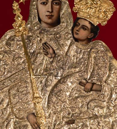 Zdjęcie nr 2: Obraz Matki Boskiej z Dzieciątkiem w kształcie stojącego prostokąta zamkniętego łukiem nadwieszonym. W centrum przedstawiona została Maria, trzymająca na lewej ręce Dzieciątko Jezus, z berłem w prawej. Maria spogląda na widza, ma owalną twarz o dużych, brązowych oczach, wąskim nosie i niewielkich, kształtnych ustach. Dzieciątko patrzy przed siebie, prawą ręką czyni gest błogosławieństwa, lewą trzyma rękę Marii. Poza widocznymi twarzami, rękami oraz stopami Dzieciątka resztę postaci zakrywają srebrne sukienki i złocone korony. Postacie zostały umieszczone na tle wykonanym z czerwonej tkaniny. Sukienki pokrywa gęsty wzór liściasto-kwiatowy złożony z rozwiniętych kwiatów i bujnych, mięsistych liści, z szerokim lamowaniem zdobionym rombowymi kaboszonami i krzyżykami. Dolna partia jest nowsza, dosztukowana i pokryta podobnym, choć rzadszym wzorem. Korony są złocone, wysokie, wypukłe, częściowo puklowane i trybowane w ornament z bujnej wici akantowej, nad czołem zdobione szlakiem z kaboszonów, a u  góry perełkowaniem i zwieńczone kulą z krzyżem. Wokół głowy Marii dodatkowo 12 gwiazdek. Matka Boska w prawej dłoni trzyma berło o rękojeści zdobionej dołem liśćmi akantu, a u góry rombowymi kaboszonami. 