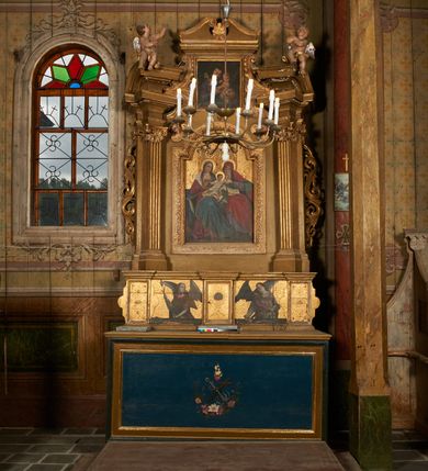 Zdjęcie nr 11: Predella ustawiona na niskim, profilowanym cokole, zamknięta profilowanym gzymsem, z wyodrębnionymi skrajnie dwoma pilastrami oraz tabernakulum po środku. Pilastry wypełnione tłoczonym w zaprawie i złoconym ornamentem okuciowym z kaboszonami i rautami, flankowane uszami w formie łuku półkolistego nadwieszonego z uskokiem. Drzwiczki tabernakulum w formie stojącego prostokąta, ujęte profilowaną ramą, w polu przedstawienie kielicha z hostią w glorii promienistej. W polach pomiędzy pilastrami a tabernakulum malowane przedstawienia aniołów z arma Christi przed parapetem, na złoconym i tłoczonym w zaprawie tle wypełnionym ornamentem roślinnym z elementami rozet, wici roślinnej i lilii heraldycznych. W lewej części półpostać anioła zwróconego w trzech czwartych w prawo, z uniesioną głową skierowaną w przeciwną stronę. W prawej, uniesionej ręce trzyma trzcinę, lewą opuszczoną wskazuje na parapet, gdzie leżą korona cierniowa, łańcuchy i sznur. Anioł ma owalną twarz, o wąskich, migdałowatych oczach skierowanych w górę, wąskim nosie i pełnych, rozchylonych ustach, okoloną rozwianymi, jasnobrązowymi włosami, sięgającymi ramion. Ubrany jest w szarą szatę spodnią z długimi, wąskimi rękawami i zieloną szatę wierzchnią z szerokimi rękawami i czerwonym kołnierzykiem. Skrzydła ma duże, złożone. W prawej części półpostać anioła ukazanego frontalnie z głową skierowaną w prawo. Lewą ugiętą ręką przytrzymuje włócznię, w dłoni trzyma sznur oraz palcem wskazuje na parapet, na którym leży nóż, prawą ręka trzyma sakwę pieniędzy leżącą na parapecie. Twarz ma owalną, o wąskich, migdałowatych oczach, prostym nosie i pełnych ustach, okoloną falowanymi włosami, sięgającymi ramion. Anioł ubrany jest w biało-szarą szatę spodnią o wąskich rękawach oraz ciemnoróżowy płaszcz skrzyżowany na piersi. Skrzydła duże, złożone.