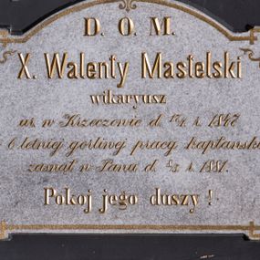 Zdjęcie nr 1: Tablica epitafijna w kształcie leżącego prostokąta zamkniętego nadwieszonym łukiem odcinkowym, ujęta prostym, drewnianym obramieniem z uszakami malowanym na czarno, na osi, w dolnej części zdobionym dekoracyjnym podwieszeniem przypominającym uproszczoną formę stykających się wolut. Dodatkowo tablica epitafium ujęta jest malowanym, złotym obramieniem z motywem lilijki w zwieńczeniu, krzyżami w narożach górnych oraz palmetkami w narożach dolnych. Pośrodku tablicy złocona inskrypcja w języku polskim „D(EO) O(PTIMO) M(AXIMO) / X(iądz) Walenty Mastelski / wikaryusz / ur(odzony) w Krzeczowie d(nia) 17./1. r(oku) 1847 / po 6 letniej gorliwej pracy kapłanskiej / zasnął w Panu d(nia) 8./5. r(oku) 1881. / Pokoj jego duszy!”.  