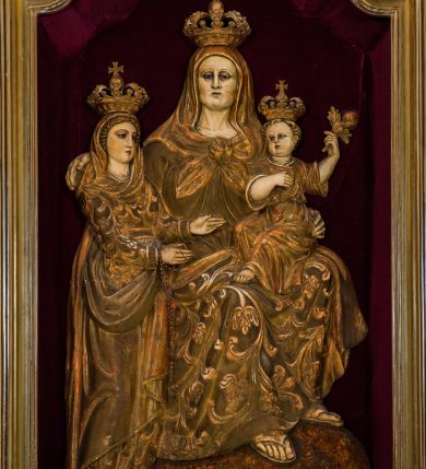 Zdjęcie nr 1: Płaskorzeźba przedstawia całopostaciowy wizerunek św. Anny Samotrzeć. Ukazana w pozycji siedzącej święta zwrócona jest w trzech czwartych w prawo, z głową skierowaną na wprost. Owalną twarz charakteryzują wyraziste, surowe rysy dojrzałej kobiety. Strój św. Anny składa się z sukni, osłaniającego nogi płaszcza zdobionego stylizowaną ornamentyką roślinną oraz osłaniającej głowę chusty związanej na piersiach. Na stopach są sandały. Święta lewą ręką podtrzymuje siedzące na jej kolanach Dzieciątko Jezus zwrócone w trzech czwartych w prawo. Zaokrągloną twarz charakteryzują pełne, dziecięce rysy, włosy są krótkie skręcone. Jezus ubrany jest w długą, przewiązaną w pasie suknię z podwiniętymi rękawami. Prawą rękę wspiera na piersiach, lewą unosi, trzymając w dłoni gałązkę różaną. Po lewej stronie, obok św. Anny stoi Maria ukazana jako kilkunastoletnia dziewczyna. Święta Anna wspiera prawą rękę na prawym ramieniu córki. Maria zwrócona jest w trzech czwartych w lewo. Owalną twarz charakteryzują młodzieńcze rysy. Ubrana jest w zdobioną ornamentem roślinnym suknię oraz osłaniający nogi, podpięty w pasie płaszcz. Na głowę ma nałożoną opadającą na ramiona chustę, odsłaniającą częściowo włosy. Maria wyciąga ręce w kierunku Dzieciątka Jezus. Na głowach postaci znajdują się zamknięte korony. Odsłonięte partie ciała utrzymane są w bladym odcieniu karnacji. Ubiór oraz atrybuty są złocone. Płaskorzeźba umieszczona jest w płytkiej niszy w kształcie stojącego prostokąta zamkniętego uskokowym łukiem nadwieszonym, wyłożonej bordowym aksamitem oraz ujętej profilowaną, złoconą ramą.    