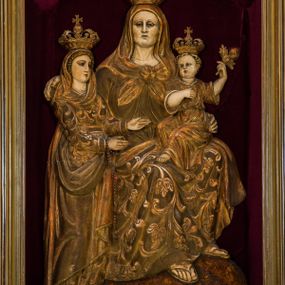 Zdjęcie nr 1: Płaskorzeźba przedstawia całopostaciowy wizerunek św. Anny Samotrzeć. Ukazana w pozycji siedzącej święta zwrócona jest w trzech czwartych w prawo, z głową skierowaną na wprost. Owalną twarz charakteryzują wyraziste, surowe rysy dojrzałej kobiety. Strój św. Anny składa się z sukni, osłaniającego nogi płaszcza zdobionego stylizowaną ornamentyką roślinną oraz osłaniającej głowę chusty związanej na piersiach. Na stopach są sandały. Święta lewą ręką podtrzymuje siedzące na jej kolanach Dzieciątko Jezus zwrócone w trzech czwartych w prawo. Zaokrągloną twarz charakteryzują pełne, dziecięce rysy, włosy są krótkie skręcone. Jezus ubrany jest w długą, przewiązaną w pasie suknię z podwiniętymi rękawami. Prawą rękę wspiera na piersiach, lewą unosi, trzymając w dłoni gałązkę różaną. Po lewej stronie, obok św. Anny stoi Maria ukazana jako kilkunastoletnia dziewczyna. Święta Anna wspiera prawą rękę na prawym ramieniu córki. Maria zwrócona jest w trzech czwartych w lewo. Owalną twarz charakteryzują młodzieńcze rysy. Ubrana jest w zdobioną ornamentem roślinnym suknię oraz osłaniający nogi, podpięty w pasie płaszcz. Na głowę ma nałożoną opadającą na ramiona chustę, odsłaniającą częściowo włosy. Maria wyciąga ręce w kierunku Dzieciątka Jezus. Na głowach postaci znajdują się zamknięte korony. Odsłonięte partie ciała utrzymane są w bladym odcieniu karnacji. Ubiór oraz atrybuty są złocone. Płaskorzeźba umieszczona jest w płytkiej niszy w kształcie stojącego prostokąta zamkniętego uskokowym łukiem nadwieszonym, wyłożonej bordowym aksamitem oraz ujętej profilowaną, złoconą ramą.    