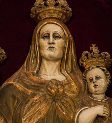 Zdjęcie nr 5: Płaskorzeźba przedstawia całopostaciowy wizerunek św. Anny Samotrzeć. Ukazana w pozycji siedzącej święta zwrócona jest w trzech czwartych w prawo, z głową skierowaną na wprost. Owalną twarz charakteryzują wyraziste, surowe rysy dojrzałej kobiety. Strój św. Anny składa się z sukni, osłaniającego nogi płaszcza zdobionego stylizowaną ornamentyką roślinną oraz osłaniającej głowę chusty związanej na piersiach. Na stopach są sandały. Święta lewą ręką podtrzymuje siedzące na jej kolanach Dzieciątko Jezus zwrócone w trzech czwartych w prawo. Zaokrągloną twarz charakteryzują pełne, dziecięce rysy, włosy są krótkie skręcone. Jezus ubrany jest w długą, przewiązaną w pasie suknię z podwiniętymi rękawami. Prawą rękę wspiera na piersiach, lewą unosi, trzymając w dłoni gałązkę różaną. Po lewej stronie, obok św. Anny stoi Maria ukazana jako kilkunastoletnia dziewczyna. Święta Anna wspiera prawą rękę na prawym ramieniu córki. Maria zwrócona jest w trzech czwartych w lewo. Owalną twarz charakteryzują młodzieńcze rysy. Ubrana jest w zdobioną ornamentem roślinnym suknię oraz osłaniający nogi, podpięty w pasie płaszcz. Na głowę ma nałożoną opadającą na ramiona chustę, odsłaniającą częściowo włosy. Maria wyciąga ręce w kierunku Dzieciątka Jezus. Na głowach postaci znajdują się zamknięte korony. Odsłonięte partie ciała utrzymane są w bladym odcieniu karnacji. Ubiór oraz atrybuty są złocone. Płaskorzeźba umieszczona jest w płytkiej niszy w kształcie stojącego prostokąta zamkniętego uskokowym łukiem nadwieszonym, wyłożonej bordowym aksamitem oraz ujętej profilowaną, złoconą ramą.    