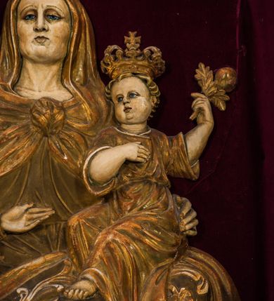 Zdjęcie nr 6: Płaskorzeźba przedstawia całopostaciowy wizerunek św. Anny Samotrzeć. Ukazana w pozycji siedzącej święta zwrócona jest w trzech czwartych w prawo, z głową skierowaną na wprost. Owalną twarz charakteryzują wyraziste, surowe rysy dojrzałej kobiety. Strój św. Anny składa się z sukni, osłaniającego nogi płaszcza zdobionego stylizowaną ornamentyką roślinną oraz osłaniającej głowę chusty związanej na piersiach. Na stopach są sandały. Święta lewą ręką podtrzymuje siedzące na jej kolanach Dzieciątko Jezus zwrócone w trzech czwartych w prawo. Zaokrągloną twarz charakteryzują pełne, dziecięce rysy, włosy są krótkie skręcone. Jezus ubrany jest w długą, przewiązaną w pasie suknię z podwiniętymi rękawami. Prawą rękę wspiera na piersiach, lewą unosi, trzymając w dłoni gałązkę różaną. Po lewej stronie, obok św. Anny stoi Maria ukazana jako kilkunastoletnia dziewczyna. Święta Anna wspiera prawą rękę na prawym ramieniu córki. Maria zwrócona jest w trzech czwartych w lewo. Owalną twarz charakteryzują młodzieńcze rysy. Ubrana jest w zdobioną ornamentem roślinnym suknię oraz osłaniający nogi, podpięty w pasie płaszcz. Na głowę ma nałożoną opadającą na ramiona chustę, odsłaniającą częściowo włosy. Maria wyciąga ręce w kierunku Dzieciątka Jezus. Na głowach postaci znajdują się zamknięte korony. Odsłonięte partie ciała utrzymane są w bladym odcieniu karnacji. Ubiór oraz atrybuty są złocone. Płaskorzeźba umieszczona jest w płytkiej niszy w kształcie stojącego prostokąta zamkniętego uskokowym łukiem nadwieszonym, wyłożonej bordowym aksamitem oraz ujętej profilowaną, złoconą ramą.    