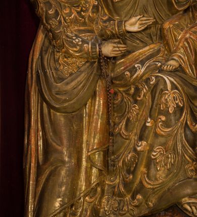 Zdjęcie nr 7: Płaskorzeźba przedstawia całopostaciowy wizerunek św. Anny Samotrzeć. Ukazana w pozycji siedzącej święta zwrócona jest w trzech czwartych w prawo, z głową skierowaną na wprost. Owalną twarz charakteryzują wyraziste, surowe rysy dojrzałej kobiety. Strój św. Anny składa się z sukni, osłaniającego nogi płaszcza zdobionego stylizowaną ornamentyką roślinną oraz osłaniającej głowę chusty związanej na piersiach. Na stopach są sandały. Święta lewą ręką podtrzymuje siedzące na jej kolanach Dzieciątko Jezus zwrócone w trzech czwartych w prawo. Zaokrągloną twarz charakteryzują pełne, dziecięce rysy, włosy są krótkie skręcone. Jezus ubrany jest w długą, przewiązaną w pasie suknię z podwiniętymi rękawami. Prawą rękę wspiera na piersiach, lewą unosi, trzymając w dłoni gałązkę różaną. Po lewej stronie, obok św. Anny stoi Maria ukazana jako kilkunastoletnia dziewczyna. Święta Anna wspiera prawą rękę na prawym ramieniu córki. Maria zwrócona jest w trzech czwartych w lewo. Owalną twarz charakteryzują młodzieńcze rysy. Ubrana jest w zdobioną ornamentem roślinnym suknię oraz osłaniający nogi, podpięty w pasie płaszcz. Na głowę ma nałożoną opadającą na ramiona chustę, odsłaniającą częściowo włosy. Maria wyciąga ręce w kierunku Dzieciątka Jezus. Na głowach postaci znajdują się zamknięte korony. Odsłonięte partie ciała utrzymane są w bladym odcieniu karnacji. Ubiór oraz atrybuty są złocone. Płaskorzeźba umieszczona jest w płytkiej niszy w kształcie stojącego prostokąta zamkniętego uskokowym łukiem nadwieszonym, wyłożonej bordowym aksamitem oraz ujętej profilowaną, złoconą ramą.    