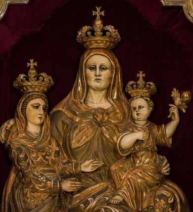 Zdjęcie nr 3: Płaskorzeźba przedstawia całopostaciowy wizerunek św. Anny Samotrzeć. Ukazana w pozycji siedzącej święta zwrócona jest w trzech czwartych w prawo, z głową skierowaną na wprost. Owalną twarz charakteryzują wyraziste, surowe rysy dojrzałej kobiety. Strój św. Anny składa się z sukni, osłaniającego nogi płaszcza zdobionego stylizowaną ornamentyką roślinną oraz osłaniającej głowę chusty związanej na piersiach. Na stopach są sandały. Święta lewą ręką podtrzymuje siedzące na jej kolanach Dzieciątko Jezus zwrócone w trzech czwartych w prawo. Zaokrągloną twarz charakteryzują pełne, dziecięce rysy, włosy są krótkie skręcone. Jezus ubrany jest w długą, przewiązaną w pasie suknię z podwiniętymi rękawami. Prawą rękę wspiera na piersiach, lewą unosi, trzymając w dłoni gałązkę różaną. Po lewej stronie, obok św. Anny stoi Maria ukazana jako kilkunastoletnia dziewczyna. Święta Anna wspiera prawą rękę na prawym ramieniu córki. Maria zwrócona jest w trzech czwartych w lewo. Owalną twarz charakteryzują młodzieńcze rysy. Ubrana jest w zdobioną ornamentem roślinnym suknię oraz osłaniający nogi, podpięty w pasie płaszcz. Na głowę ma nałożoną opadającą na ramiona chustę, odsłaniającą częściowo włosy. Maria wyciąga ręce w kierunku Dzieciątka Jezus. Na głowach postaci znajdują się zamknięte korony. Odsłonięte partie ciała utrzymane są w bladym odcieniu karnacji. Ubiór oraz atrybuty są złocone. Płaskorzeźba umieszczona jest w płytkiej niszy w kształcie stojącego prostokąta zamkniętego uskokowym łukiem nadwieszonym, wyłożonej bordowym aksamitem oraz ujętej profilowaną, złoconą ramą.    