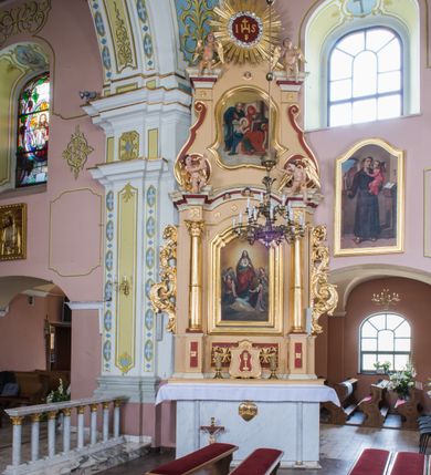 Zdjęcie nr 2: Obraz uzyskał kształt stojącego prostokąta ujętego profilowaną i złoconą ramą ołtarza, zamkniętą u góry łukiem odcinkowym, nadwieszonym. W centrum kompozycji wyobrażono frontalnie stojącego na obłokach Chrystusa ukazującego swoje serce, adorowanego przez anioły. Zbawiciel ma lekko pochyloną głowę w lewo. Prawą rękę unosi w geście błogosławieństwa, lewą zaś wskazuje na płonące na środku piersi serce. Obie dłonie są przebite. Wyobrażone na piersi serce na tle jasnych promieni otacza korona cierniowa. Z rany oraz u góry serca widać wydobywające się płomienie. Twarz Chrystusa jest pociągła. Otaczają je długie brązowe włosy opadające puklami na ramiona oraz brązowa broda. Twarz Zbawiciela charakteryzuje się wysokim czołem, długim, wąskim nosem i wydatnymi ustami. Chrystus szeroko otwartymi ciemnymi oczami wpatruje się w widza. Jego głowę otacza złocista poświata. Ubrany jest w luźną, turkusową tunikę przewiązaną w pasie i narzucony na ramiona czerwony płaszcz podbity ciemną ochrą, którego prawa poła przerzucona jest przez lewe przedramię. Zbawiciel został ukazany na jasnoochrowym tle. Po obu stronach na obłokach widać czterech adorujących Jezusa uskrzydlonych aniołów. Dwaj z przodu klęczą, a dwaj z tyłu stoją. Ubrani są w luźne szaty w pastelowych, przygaszonych kolorach.