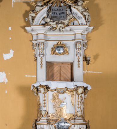 Zdjęcie nr 2: Ambona zawieszona jest na ścianie, z wejściem w zaplecku. Kosz ambony, na rzucie owalu, artykułują cztery pilastry osadzone na obiegającym jego podstawę cokole, wspierające wyłamujący się ponad nimi gzyms wieńczący. Podwieszenie kosza jest w formie czterech wolutowych kabłąków spiętych spłaszczonym pierścieniem. Zaplecek jest w formie prostokątnej płyciny ujętej parą pilastrów wspierających wyłamujące się ponad nimi belkowanie z wydatnym gzymsem. Zwieńczenie ma formę półkolistego przyczółka. Pola kosza ambony wypełniają płaskorzeźbione przedstawienia trzech świętych Janów. W środkowym znajduje się św. Jan Kanty siedzący przy pulpicie z otwartą księgą, poniżej znajduje się

kartusz herbowy, o krawędziach zdobionych ornamentyką akantową, z napisem: „Et lex / clementia / in linqua / eius”, natomiast w medalionie powyżej jest zamknięta księga z siedmioma pieczęciami. Po lewej znajduje się św. Jan Ewangelista, poniżej na kartuszu napis: „Quia verum est testimo/nium eius”, w medalionie zaś klatka. Po prawej jest przedstawienie św. Jana Chrzciciela z kartuszem z napisem: „Et enim / manus Domini / errat /cum / suo” oraz medalionem ze znakiem ręki trzymającej trąbę. Pilastry zdobią motywy kampanulli oraz uskrzydlonych główek anielskich, z których niektóre są na tle konch. Tuż pod gzymsem wieńczącym podwieszona jest girlanda kwiatowa. Belkowanie zaplecka ambony wsparte jest dodatkowo na wolutowych wspornikach, trzony pilastrów zdobią kampanulle. Po środku płyciny zaplecka, nad wejściem znajduje się ujęty akantem kartusz herbowy z napisem: „Docebit vos omnia”. Zwieńczenie ambony zdobią figurki dwóch par aniołków, z czego jedna usytuowana jest na krawędziach przyczółka, druga wypełnia pole tympanonu. Ukazane naprzeciw siebie w pozycjach półleżących, o przeplatających się nogach obuczącz podtrzymują otwartą księgę z napisem: „EVANGELIUM IESU CHRISTI”. Szczyt zwieńczenia jest w formie promienistej glorii z gołębicą Ducha Świętego po środku. Struktura ambony jest pomalowana na biało, podobnie, jak odsłonięte partie ciał aniołków. Detal architektoniczny, ornamnetyka, szaty i skrzydła są złocone i srebrzone.