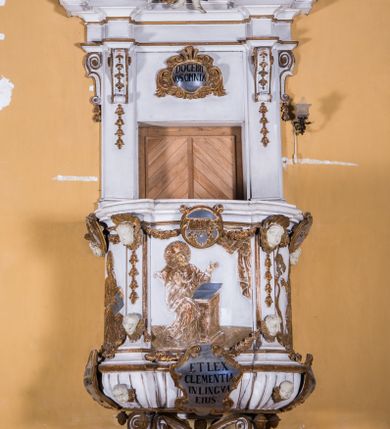 Zdjęcie nr 1: Ambona zawieszona jest na ścianie, z wejściem w zaplecku. Kosz ambony, na rzucie owalu, artykułują cztery pilastry osadzone na obiegającym jego podstawę cokole, wspierające wyłamujący się ponad nimi gzyms wieńczący. Podwieszenie kosza jest w formie czterech wolutowych kabłąków spiętych spłaszczonym pierścieniem. Zaplecek jest w formie prostokątnej płyciny ujętej parą pilastrów wspierających wyłamujące się ponad nimi belkowanie z wydatnym gzymsem. Zwieńczenie ma formę półkolistego przyczółka. Pola kosza ambony wypełniają płaskorzeźbione przedstawienia trzech świętych Janów. W środkowym znajduje się św. Jan Kanty siedzący przy pulpicie z otwartą księgą, poniżej znajduje się

kartusz herbowy, o krawędziach zdobionych ornamentyką akantową, z napisem: „Et lex / clementia / in linqua / eius”, natomiast w medalionie powyżej jest zamknięta księga z siedmioma pieczęciami. Po lewej znajduje się św. Jan Ewangelista, poniżej na kartuszu napis: „Quia verum est testimo/nium eius”, w medalionie zaś klatka. Po prawej jest przedstawienie św. Jana Chrzciciela z kartuszem z napisem: „Et enim / manus Domini / errat /cum / suo” oraz medalionem ze znakiem ręki trzymającej trąbę. Pilastry zdobią motywy kampanulli oraz uskrzydlonych główek anielskich, z których niektóre są na tle konch. Tuż pod gzymsem wieńczącym podwieszona jest girlanda kwiatowa. Belkowanie zaplecka ambony wsparte jest dodatkowo na wolutowych wspornikach, trzony pilastrów zdobią kampanulle. Po środku płyciny zaplecka, nad wejściem znajduje się ujęty akantem kartusz herbowy z napisem: „Docebit vos omnia”. Zwieńczenie ambony zdobią figurki dwóch par aniołków, z czego jedna usytuowana jest na krawędziach przyczółka, druga wypełnia pole tympanonu. Ukazane naprzeciw siebie w pozycjach półleżących, o przeplatających się nogach obuczącz podtrzymują otwartą księgę z napisem: „EVANGELIUM IESU CHRISTI”. Szczyt zwieńczenia jest w formie promienistej glorii z gołębicą Ducha Świętego po środku. Struktura ambony jest pomalowana na biało, podobnie, jak odsłonięte partie ciał aniołków. Detal architektoniczny, ornamnetyka, szaty i skrzydła są złocone i srebrzone.