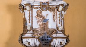 Zdjęcie nr 1: Ambona zawieszona jest na ścianie, z wejściem w zaplecku. Kosz ambony, na rzucie owalu, artykułują cztery pilastry osadzone na obiegającym jego podstawę cokole, wspierające wyłamujący się ponad nimi gzyms wieńczący. Podwieszenie kosza jest w formie czterech wolutowych kabłąków spiętych spłaszczonym pierścieniem. Zaplecek jest w formie prostokątnej płyciny ujętej parą pilastrów wspierających wyłamujące się ponad nimi belkowanie z wydatnym gzymsem. Zwieńczenie ma formę półkolistego przyczółka. Pola kosza ambony wypełniają płaskorzeźbione przedstawienia trzech świętych Janów. W środkowym znajduje się św. Jan Kanty siedzący przy pulpicie z otwartą księgą, poniżej znajduje się

kartusz herbowy, o krawędziach zdobionych ornamentyką akantową, z napisem: „Et lex / clementia / in linqua / eius”, natomiast w medalionie powyżej jest zamknięta księga z siedmioma pieczęciami. Po lewej znajduje się św. Jan Ewangelista, poniżej na kartuszu napis: „Quia verum est testimo/nium eius”, w medalionie zaś klatka. Po prawej jest przedstawienie św. Jana Chrzciciela z kartuszem z napisem: „Et enim / manus Domini / errat /cum / suo” oraz medalionem ze znakiem ręki trzymającej trąbę. Pilastry zdobią motywy kampanulli oraz uskrzydlonych główek anielskich, z których niektóre są na tle konch. Tuż pod gzymsem wieńczącym podwieszona jest girlanda kwiatowa. Belkowanie zaplecka ambony wsparte jest dodatkowo na wolutowych wspornikach, trzony pilastrów zdobią kampanulle. Po środku płyciny zaplecka, nad wejściem znajduje się ujęty akantem kartusz herbowy z napisem: „Docebit vos omnia”. Zwieńczenie ambony zdobią figurki dwóch par aniołków, z czego jedna usytuowana jest na krawędziach przyczółka, druga wypełnia pole tympanonu. Ukazane naprzeciw siebie w pozycjach półleżących, o przeplatających się nogach obuczącz podtrzymują otwartą księgę z napisem: „EVANGELIUM IESU CHRISTI”. Szczyt zwieńczenia jest w formie promienistej glorii z gołębicą Ducha Świętego po środku. Struktura ambony jest pomalowana na biało, podobnie, jak odsłonięte partie ciał aniołków. Detal architektoniczny, ornamnetyka, szaty i skrzydła są złocone i srebrzone.