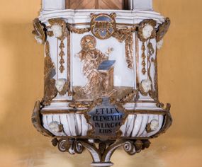 Zdjęcie nr 1: Ambona zawieszona jest na ścianie, z wejściem w zaplecku. Kosz ambony, na rzucie owalu, artykułują cztery pilastry osadzone na obiegającym jego podstawę cokole, wspierające wyłamujący się ponad nimi gzyms wieńczący. Podwieszenie kosza jest w formie czterech wolutowych kabłąków spiętych spłaszczonym pierścieniem. Zaplecek jest w formie prostokątnej płyciny ujętej parą pilastrów wspierających wyłamujące się ponad nimi belkowanie z wydatnym gzymsem. Zwieńczenie ma formę półkolistego przyczółka. Pola kosza ambony wypełniają płaskorzeźbione przedstawienia trzech świętych Janów. W środkowym znajduje się św. Jan Kanty siedzący przy pulpicie z otwartą księgą, poniżej znajduje się

kartusz herbowy, o krawędziach zdobionych ornamentyką akantową, z napisem: „Et lex / clementia / in linqua / eius”, natomiast w medalionie powyżej jest zamknięta księga z siedmioma pieczęciami. Po lewej znajduje się św. Jan Ewangelista, poniżej na kartuszu napis: „Quia verum est testimo/nium eius”, w medalionie zaś klatka. Po prawej jest przedstawienie św. Jana Chrzciciela z kartuszem z napisem: „Et enim / manus Domini / errat /cum / suo” oraz medalionem ze znakiem ręki trzymającej trąbę. Pilastry zdobią motywy kampanulli oraz uskrzydlonych główek anielskich, z których niektóre są na tle konch. Tuż pod gzymsem wieńczącym podwieszona jest girlanda kwiatowa. Belkowanie zaplecka ambony wsparte jest dodatkowo na wolutowych wspornikach, trzony pilastrów zdobią kampanulle. Po środku płyciny zaplecka, nad wejściem znajduje się ujęty akantem kartusz herbowy z napisem: „Docebit vos omnia”. Zwieńczenie ambony zdobią figurki dwóch par aniołków, z czego jedna usytuowana jest na krawędziach przyczółka, druga wypełnia pole tympanonu. Ukazane naprzeciw siebie w pozycjach półleżących, o przeplatających się nogach obuczącz podtrzymują otwartą księgę z napisem: „EVANGELIUM IESU CHRISTI”. Szczyt zwieńczenia jest w formie promienistej glorii z gołębicą Ducha Świętego po środku. Struktura ambony jest pomalowana na biało, podobnie, jak odsłonięte partie ciał aniołków. Detal architektoniczny, ornamnetyka, szaty i skrzydła są złocone i srebrzone.