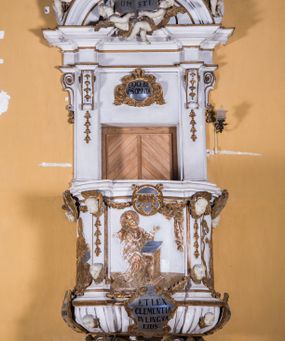 Zdjęcie nr 1: Ambona zawieszona jest na ścianie, z wejściem w zaplecku. Kosz ambony, na rzucie owalu, artykułują cztery pilastry osadzone na obiegającym jego podstawę cokole, wspierające wyłamujący się ponad nimi gzyms wieńczący. Podwieszenie kosza jest w formie czterech wolutowych kabłąków spiętych spłaszczonym pierścieniem. Zaplecek jest w formie prostokątnej płyciny ujętej parą pilastrów wspierających wyłamujące się ponad nimi belkowanie z wydatnym gzymsem. Zwieńczenie ma formę półkolistego przyczółka. Pola kosza ambony wypełniają płaskorzeźbione przedstawienia trzech świętych Janów. W środkowym znajduje się św. Jan Kanty siedzący przy pulpicie z otwartą księgą, poniżej znajduje się

kartusz herbowy, o krawędziach zdobionych ornamentyką akantową, z napisem: „Et lex / clementia / in linqua / eius”, natomiast w medalionie powyżej jest zamknięta księga z siedmioma pieczęciami. Po lewej znajduje się św. Jan Ewangelista, poniżej na kartuszu napis: „Quia verum est testimo/nium eius”, w medalionie zaś klatka. Po prawej jest przedstawienie św. Jana Chrzciciela z kartuszem z napisem: „Et enim / manus Domini / errat /cum / suo” oraz medalionem ze znakiem ręki trzymającej trąbę. Pilastry zdobią motywy kampanulli oraz uskrzydlonych główek anielskich, z których niektóre są na tle konch. Tuż pod gzymsem wieńczącym podwieszona jest girlanda kwiatowa. Belkowanie zaplecka ambony wsparte jest dodatkowo na wolutowych wspornikach, trzony pilastrów zdobią kampanulle. Po środku płyciny zaplecka, nad wejściem znajduje się ujęty akantem kartusz herbowy z napisem: „Docebit vos omnia”. Zwieńczenie ambony zdobią figurki dwóch par aniołków, z czego jedna usytuowana jest na krawędziach przyczółka, druga wypełnia pole tympanonu. Ukazane naprzeciw siebie w pozycjach półleżących, o przeplatających się nogach obuczącz podtrzymują otwartą księgę z napisem: „EVANGELIUM IESU CHRISTI”. Szczyt zwieńczenia jest w formie promienistej glorii z gołębicą Ducha Świętego po środku. Struktura ambony jest pomalowana na biało, podobnie, jak odsłonięte partie ciał aniołków. Detal architektoniczny, ornamnetyka, szaty i skrzydła są złocone i srebrzone.