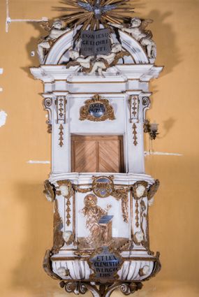 Zdjęcie nr 1: Ambona zawieszona jest na ścianie, z wejściem w zaplecku. Kosz ambony, na rzucie owalu, artykułują cztery pilastry osadzone na obiegającym jego podstawę cokole, wspierające wyłamujący się ponad nimi gzyms wieńczący. Podwieszenie kosza jest w formie czterech wolutowych kabłąków spiętych spłaszczonym pierścieniem. Zaplecek jest w formie prostokątnej płyciny ujętej parą pilastrów wspierających wyłamujące się ponad nimi belkowanie z wydatnym gzymsem. Zwieńczenie ma formę półkolistego przyczółka. Pola kosza ambony wypełniają płaskorzeźbione przedstawienia trzech świętych Janów. W środkowym znajduje się św. Jan Kanty siedzący przy pulpicie z otwartą księgą, poniżej znajduje się

kartusz herbowy, o krawędziach zdobionych ornamentyką akantową, z napisem: „Et lex / clementia / in linqua / eius”, natomiast w medalionie powyżej jest zamknięta księga z siedmioma pieczęciami. Po lewej znajduje się św. Jan Ewangelista, poniżej na kartuszu napis: „Quia verum est testimo/nium eius”, w medalionie zaś klatka. Po prawej jest przedstawienie św. Jana Chrzciciela z kartuszem z napisem: „Et enim / manus Domini / errat /cum / suo” oraz medalionem ze znakiem ręki trzymającej trąbę. Pilastry zdobią motywy kampanulli oraz uskrzydlonych główek anielskich, z których niektóre są na tle konch. Tuż pod gzymsem wieńczącym podwieszona jest girlanda kwiatowa. Belkowanie zaplecka ambony wsparte jest dodatkowo na wolutowych wspornikach, trzony pilastrów zdobią kampanulle. Po środku płyciny zaplecka, nad wejściem znajduje się ujęty akantem kartusz herbowy z napisem: „Docebit vos omnia”. Zwieńczenie ambony zdobią figurki dwóch par aniołków, z czego jedna usytuowana jest na krawędziach przyczółka, druga wypełnia pole tympanonu. Ukazane naprzeciw siebie w pozycjach półleżących, o przeplatających się nogach obuczącz podtrzymują otwartą księgę z napisem: „EVANGELIUM IESU CHRISTI”. Szczyt zwieńczenia jest w formie promienistej glorii z gołębicą Ducha Świętego po środku. Struktura ambony jest pomalowana na biało, podobnie, jak odsłonięte partie ciał aniołków. Detal architektoniczny, ornamnetyka, szaty i skrzydła są złocone i srebrzone.