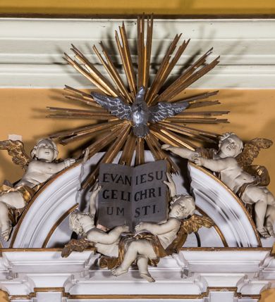 Zdjęcie nr 5: Ambona zawieszona jest na ścianie, z wejściem w zaplecku. Kosz ambony, na rzucie owalu, artykułują cztery pilastry osadzone na obiegającym jego podstawę cokole, wspierające wyłamujący się ponad nimi gzyms wieńczący. Podwieszenie kosza jest w formie czterech wolutowych kabłąków spiętych spłaszczonym pierścieniem. Zaplecek jest w formie prostokątnej płyciny ujętej parą pilastrów wspierających wyłamujące się ponad nimi belkowanie z wydatnym gzymsem. Zwieńczenie ma formę półkolistego przyczółka. Pola kosza ambony wypełniają płaskorzeźbione przedstawienia trzech świętych Janów. W środkowym znajduje się św. Jan Kanty siedzący przy pulpicie z otwartą księgą, poniżej znajduje się

kartusz herbowy, o krawędziach zdobionych ornamentyką akantową, z napisem: „Et lex / clementia / in linqua / eius”, natomiast w medalionie powyżej jest zamknięta księga z siedmioma pieczęciami. Po lewej znajduje się św. Jan Ewangelista, poniżej na kartuszu napis: „Quia verum est testimo/nium eius”, w medalionie zaś klatka. Po prawej jest przedstawienie św. Jana Chrzciciela z kartuszem z napisem: „Et enim / manus Domini / errat /cum / suo” oraz medalionem ze znakiem ręki trzymającej trąbę. Pilastry zdobią motywy kampanulli oraz uskrzydlonych główek anielskich, z których niektóre są na tle konch. Tuż pod gzymsem wieńczącym podwieszona jest girlanda kwiatowa. Belkowanie zaplecka ambony wsparte jest dodatkowo na wolutowych wspornikach, trzony pilastrów zdobią kampanulle. Po środku płyciny zaplecka, nad wejściem znajduje się ujęty akantem kartusz herbowy z napisem: „Docebit vos omnia”. Zwieńczenie ambony zdobią figurki dwóch par aniołków, z czego jedna usytuowana jest na krawędziach przyczółka, druga wypełnia pole tympanonu. Ukazane naprzeciw siebie w pozycjach półleżących, o przeplatających się nogach obuczącz podtrzymują otwartą księgę z napisem: „EVANGELIUM IESU CHRISTI”. Szczyt zwieńczenia jest w formie promienistej glorii z gołębicą Ducha Świętego po środku. Struktura ambony jest pomalowana na biało, podobnie, jak odsłonięte partie ciał aniołków. Detal architektoniczny, ornamnetyka, szaty i skrzydła są złocone i srebrzone.